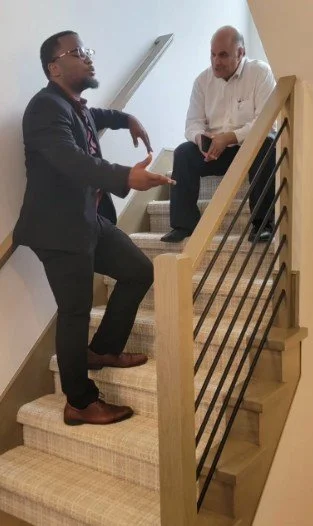 Two men talking on a staircase, one is standing and the other is sitting on the upper step.