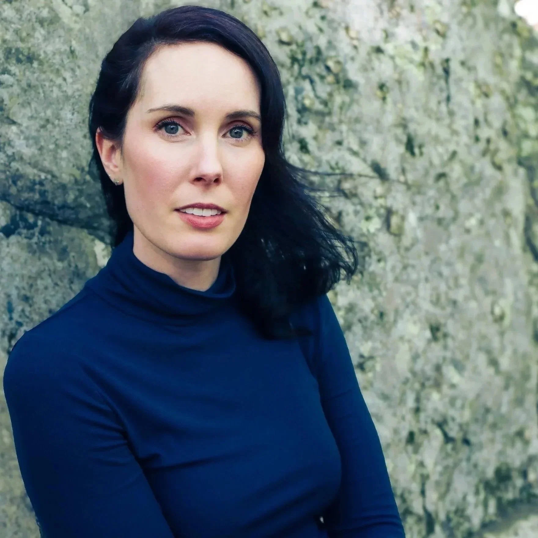 Author headshot of Evann Normandin, a woman with dark hair and blue eyes, wearing a dark blue turtleneck, standing outdoors with a blurred stone wall background.