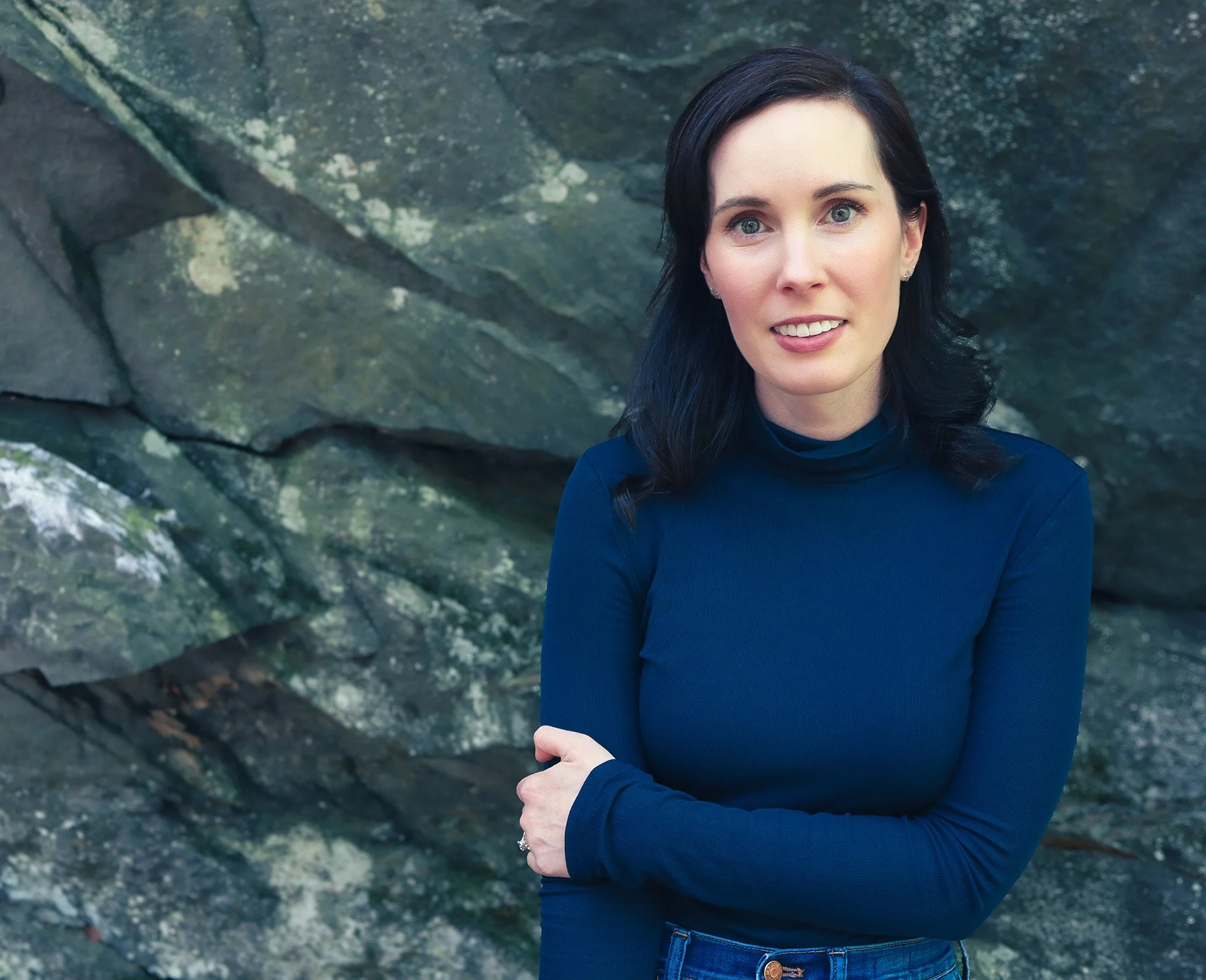 Author headshot of Evann Normandin, a woman with dark hair wearing a blue turtleneck sweater standing in front of a rocky background with her arms crossed.
