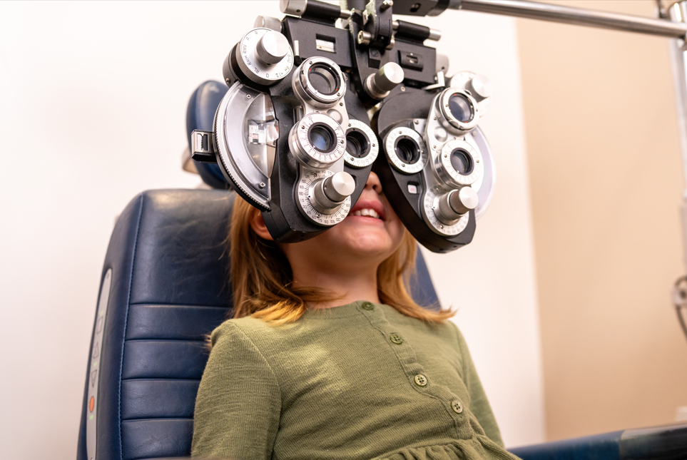 Young girl sitting in an eye exam chair while undergoing vision testing with an optometry phoropter.