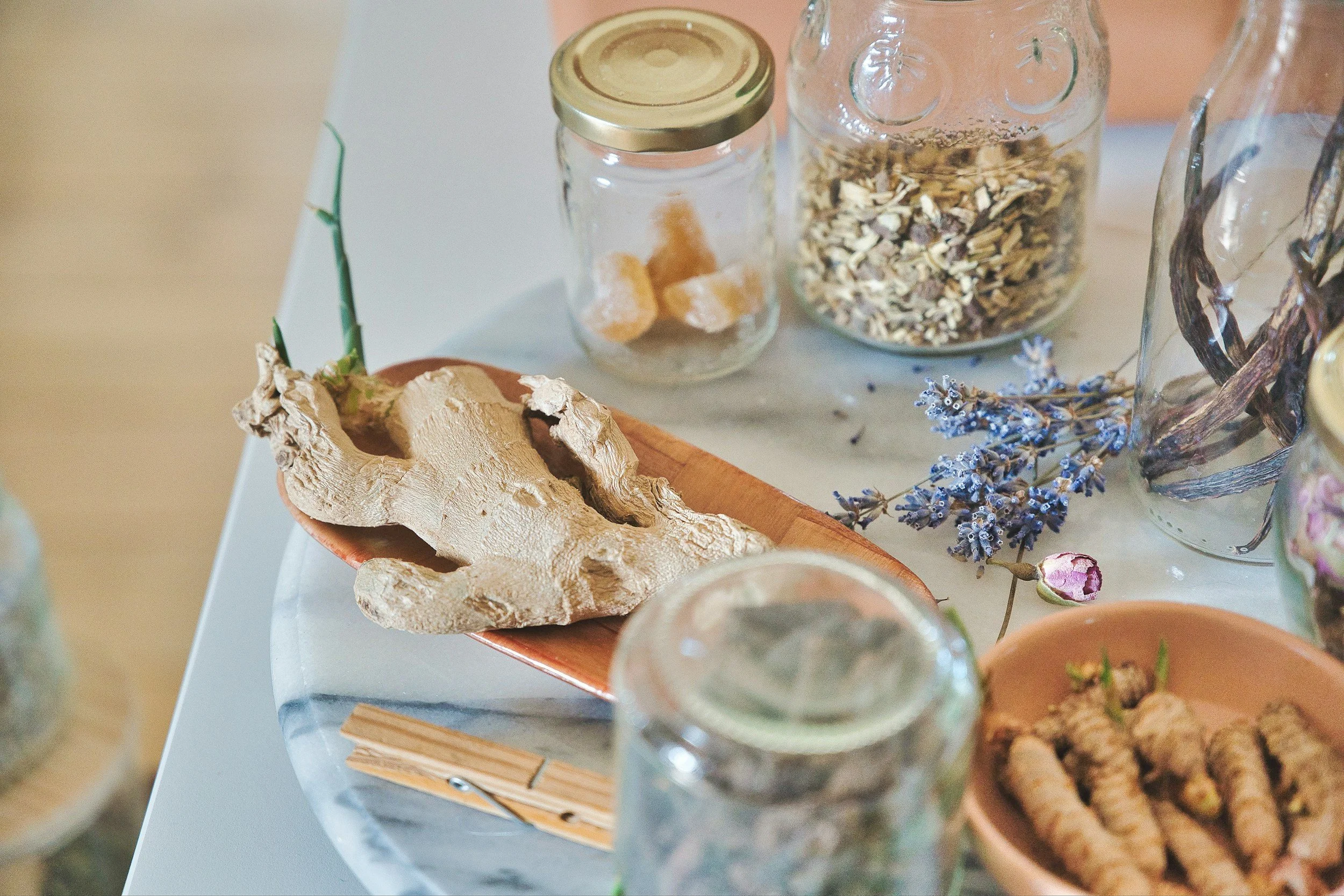 Assorted dried herbs and roots in glass jars and small dishes, some with dried flowers and spices, on a white surface.