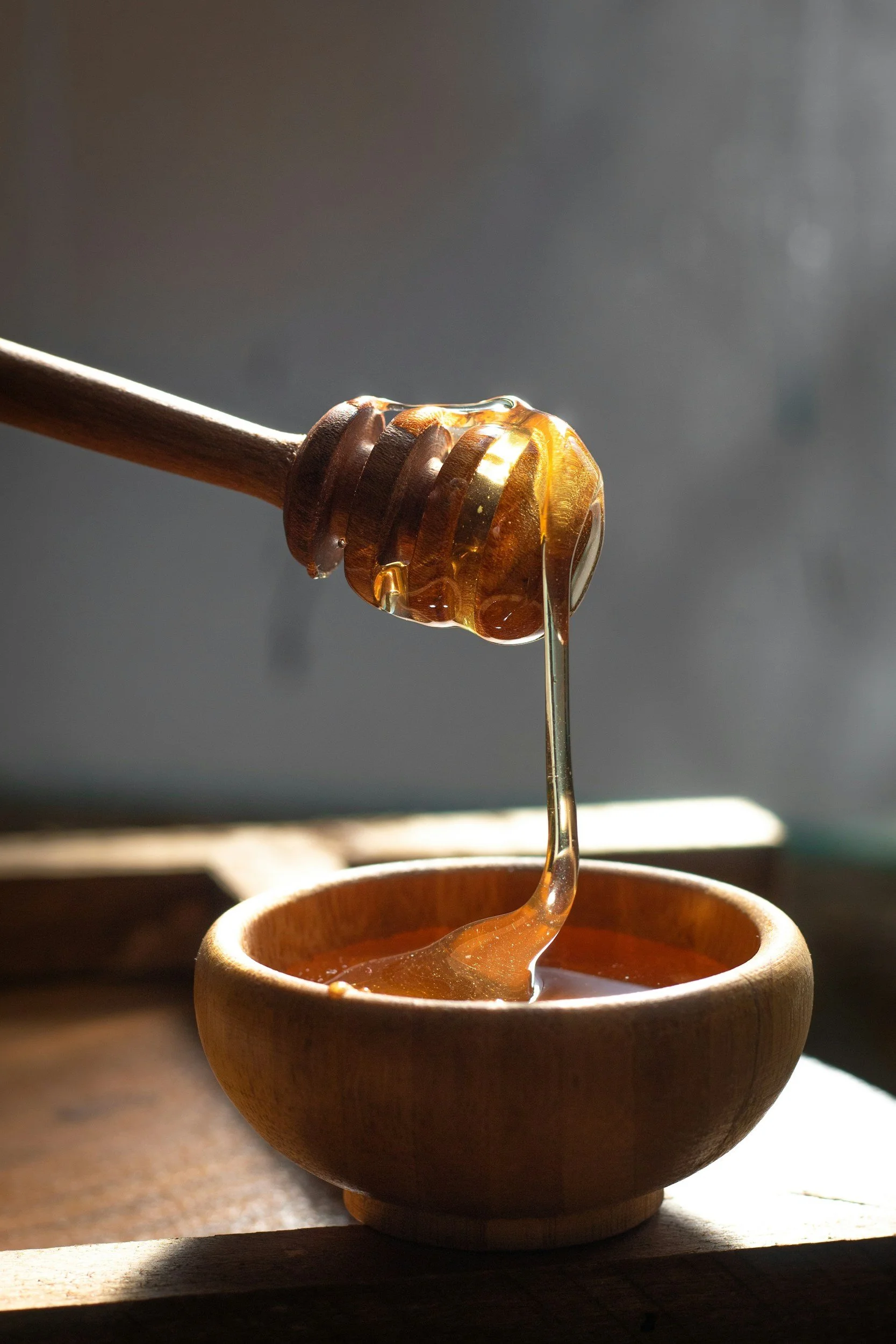 A wooden honey dipper holds dripping honey over a small wooden bowl, with sunlight illuminating the sticky, golden honey.