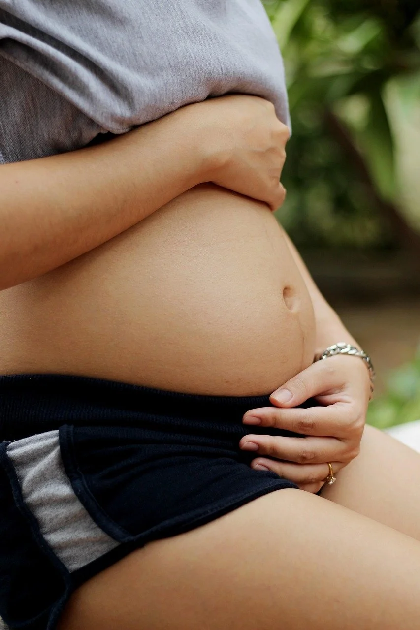 Close-up of a pregnant belly with one hand gently holding the side and the other hand resting on the lower stomach, with a blurred green background.