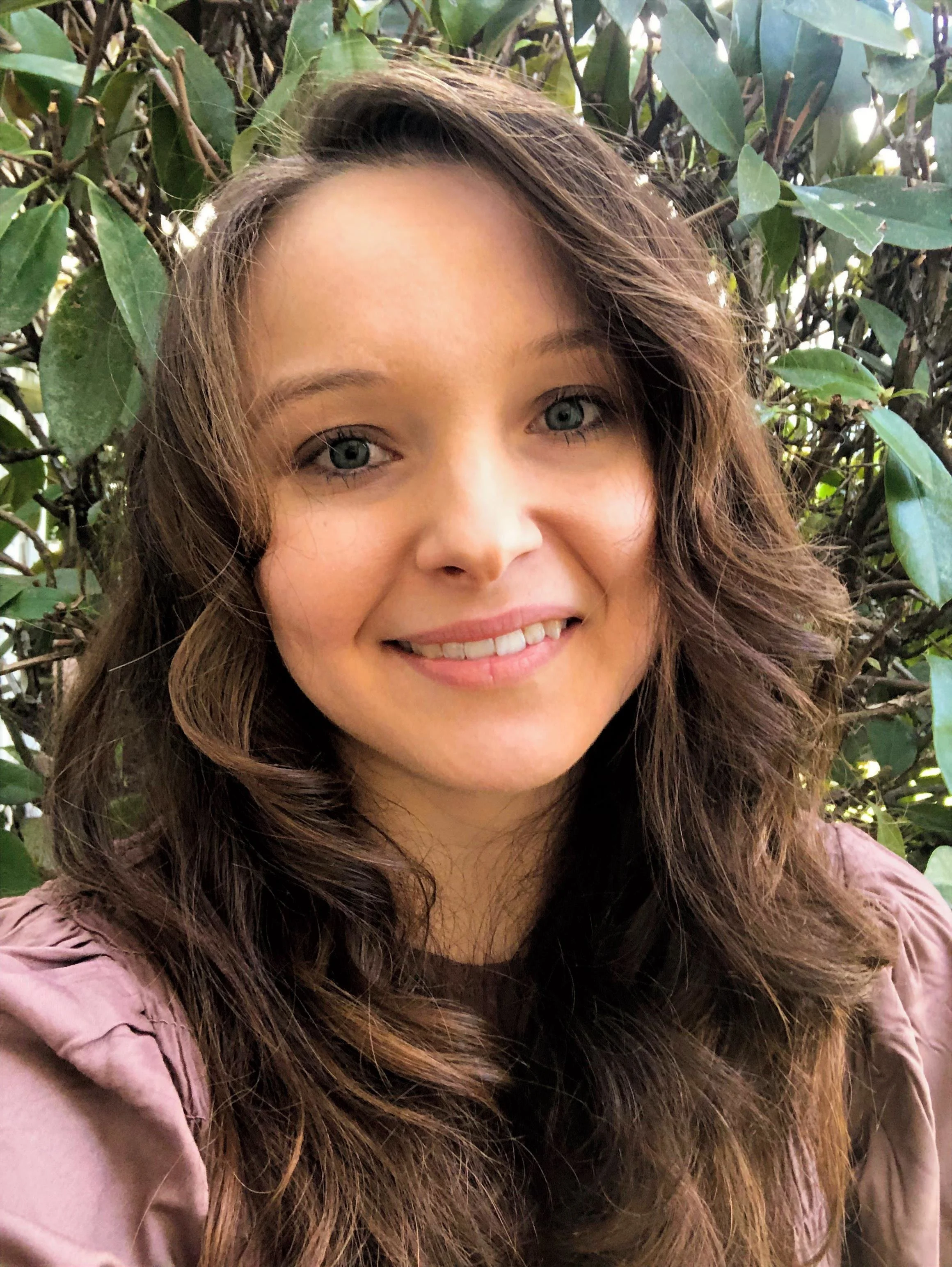 A woman with long, wavy brown hair, light skin, and blue eyes smiling, standing outdoors with green leafy foliage in the background.