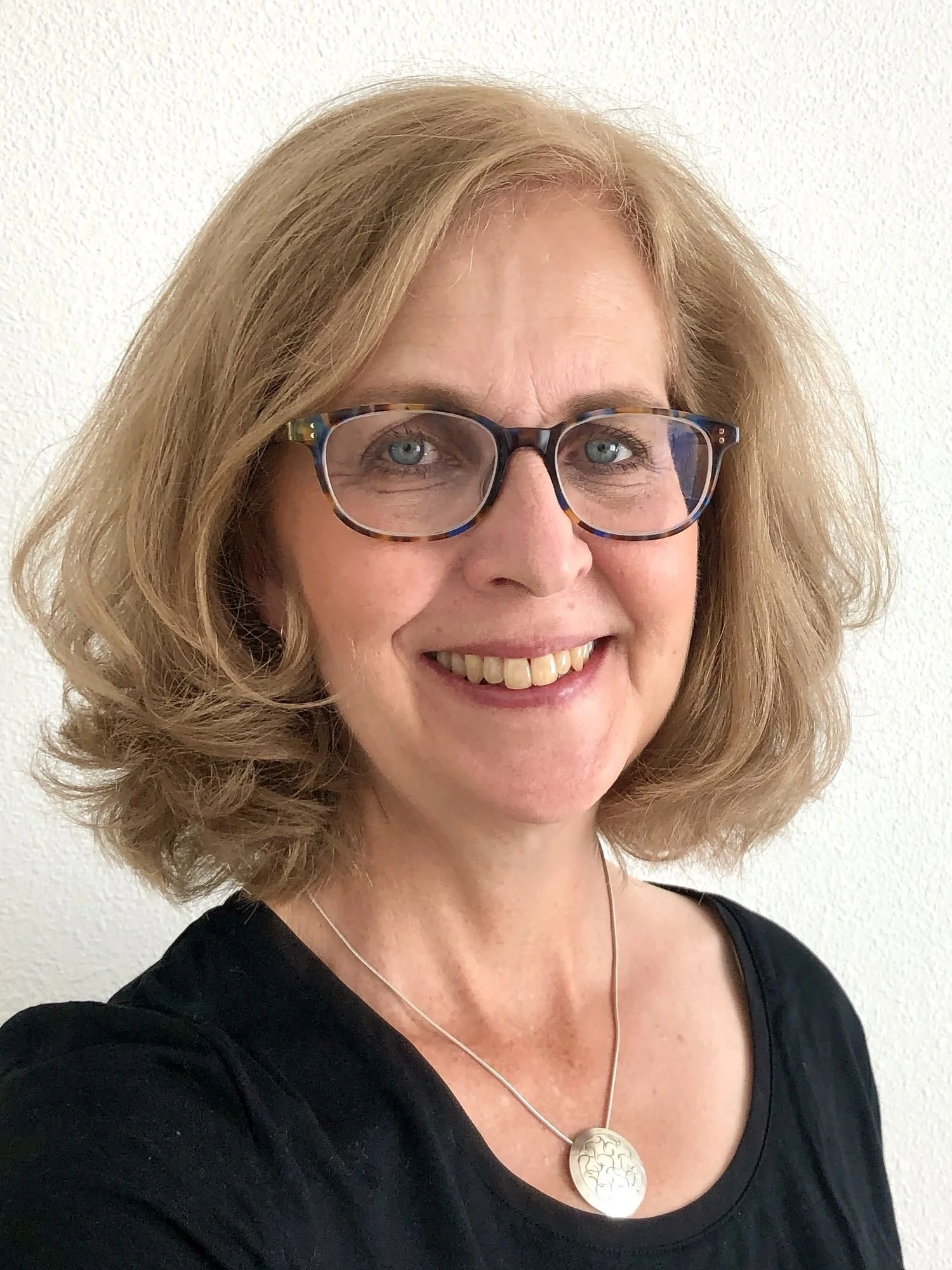 A woman with shoulder-length wavy blonde hair, wearing glasses with a tortoise shell frame, smiling, dressed in a black top, and wearing a silver necklace with a round pendant, standing against a white textured wall.