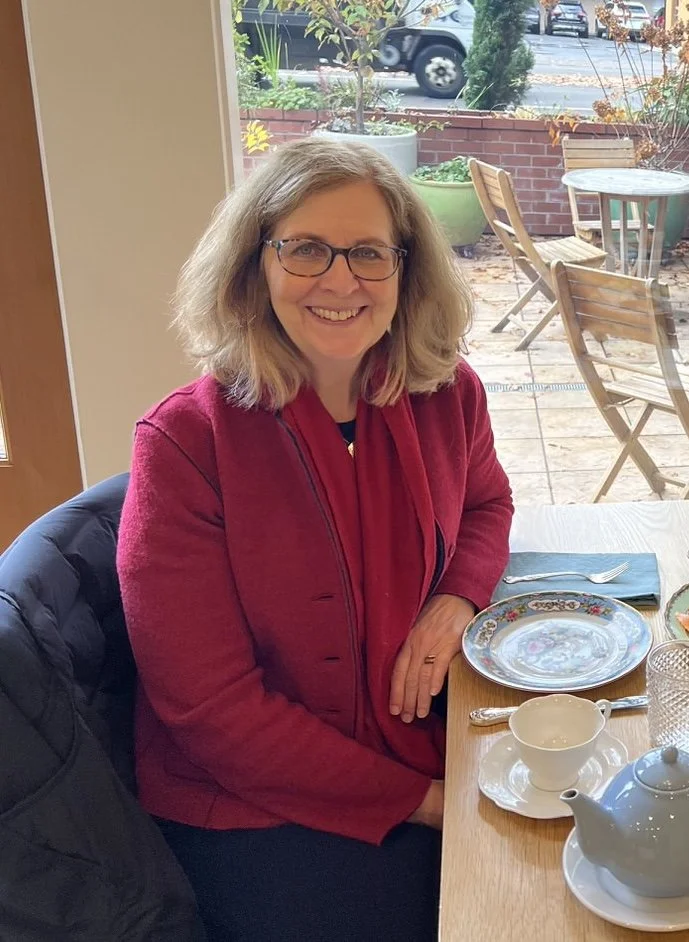 A smiling woman with glasses and shoulder-length blonde hair, wearing a red jacket, sitting at a table with a tea cup, teapot, and plates, in a cozy indoor setting near a window with a view of an outdoor patio and parked cars.