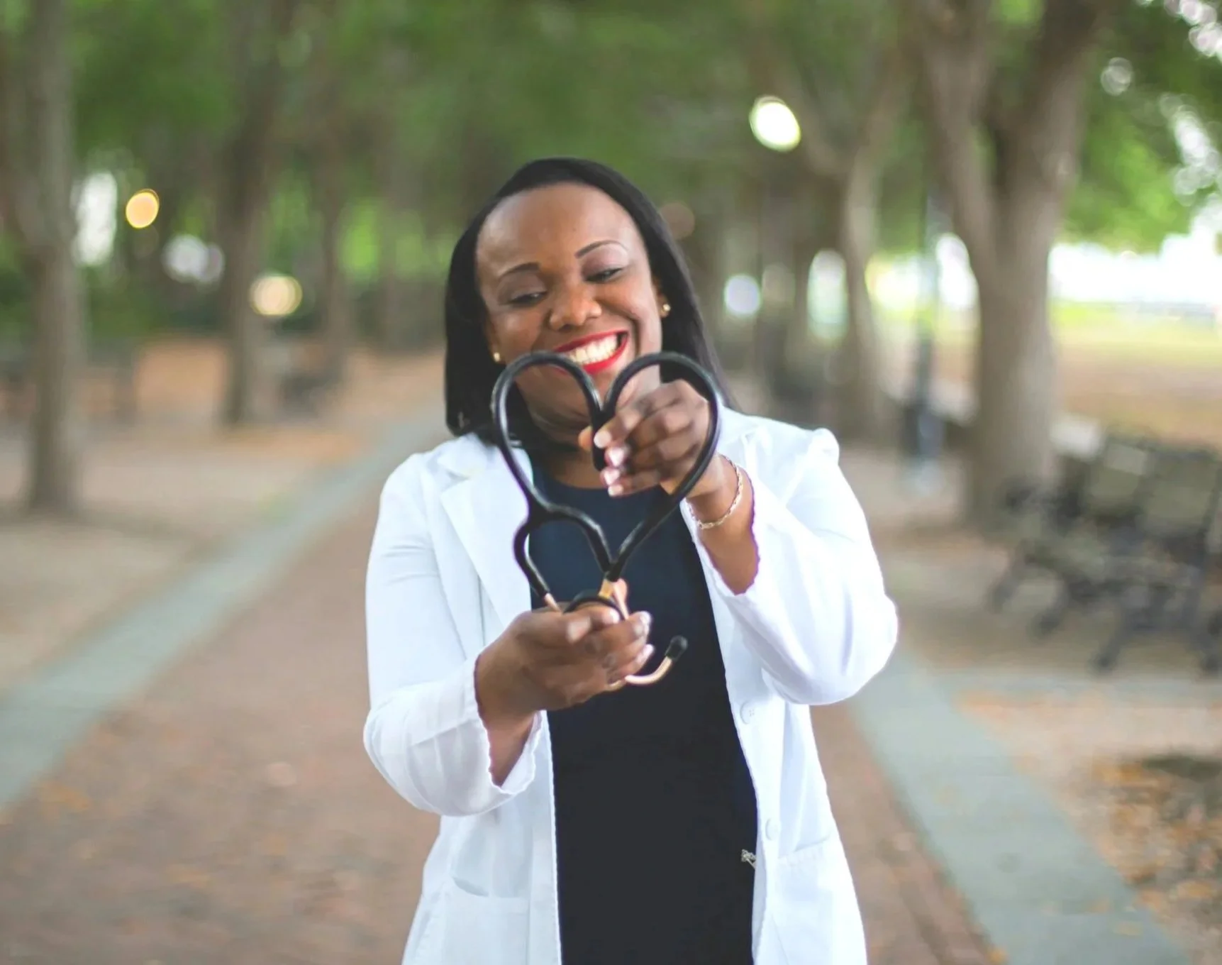 Woman physician in white medical coat, smiling with joy and energy, holding a heart-shaped stethoscope outdoors in a park.
