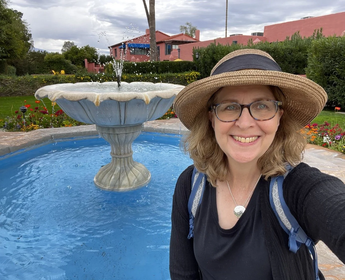 A joyful looking woman wearing a hat and backpack, sitting in front of a fountain in a garden with colorful flowers and a pink building in the background.