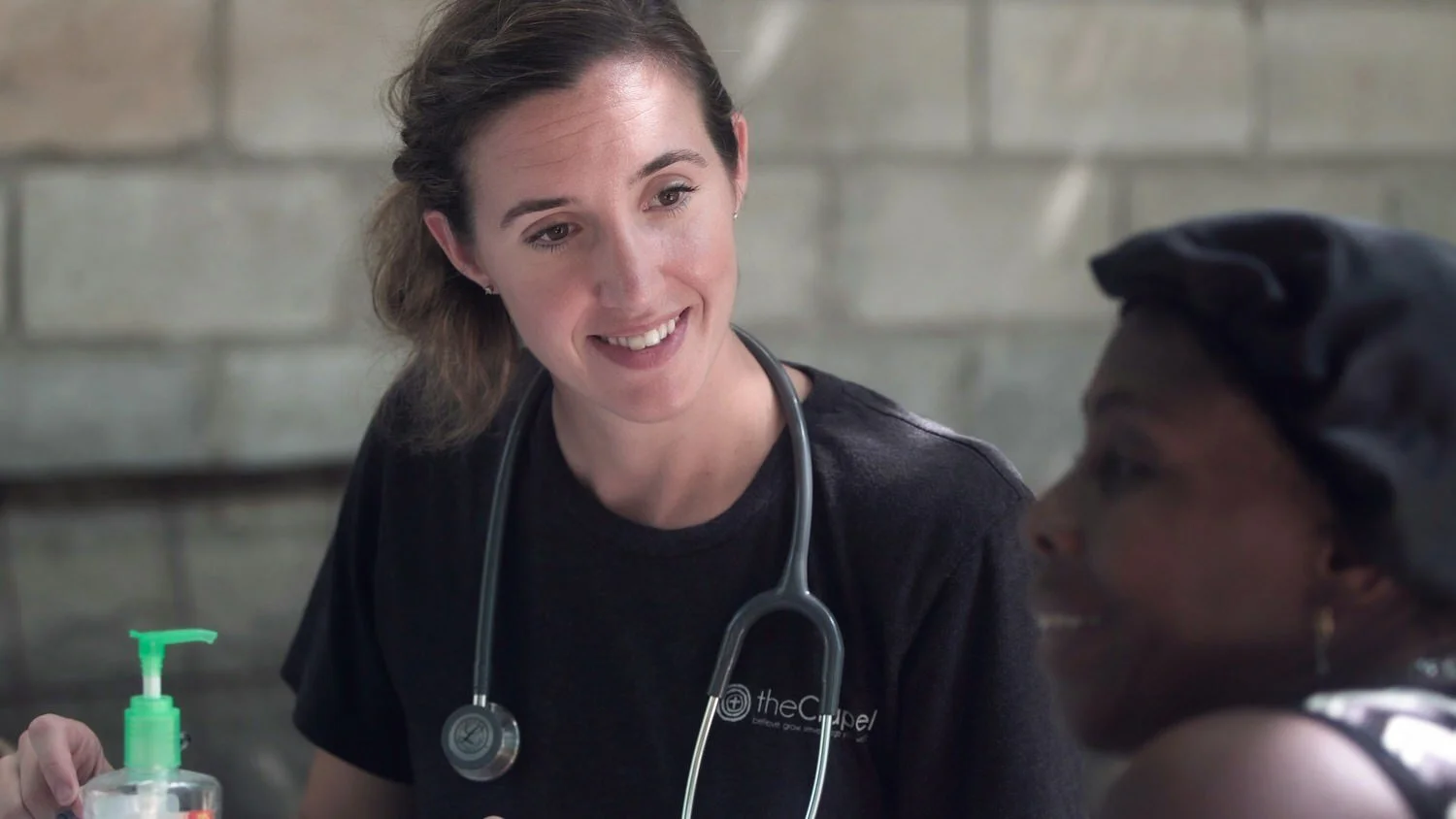 Woman physician with a stethoscope around her neck, smiling and talking to a patient while leaning in with eye contact. The patient is out of focus.