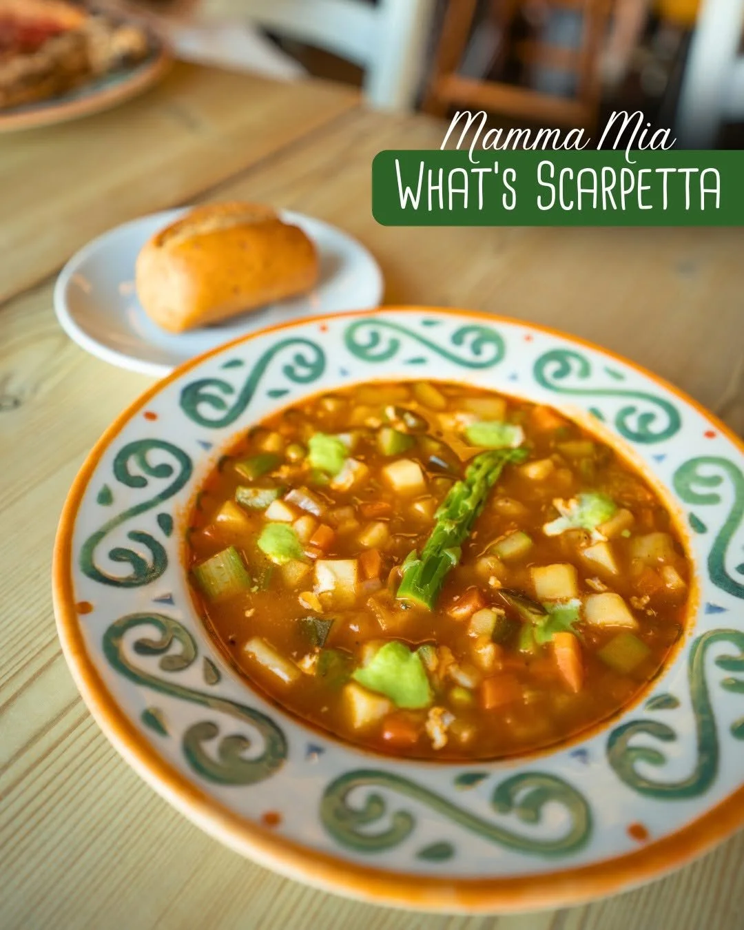 A bowl of vegetable soup with a piece of green onion on top, on a colorful decorative plate, with a roll on a small white plate in the background, and text 'Mamma Mia What's Scarpetta' on the image.