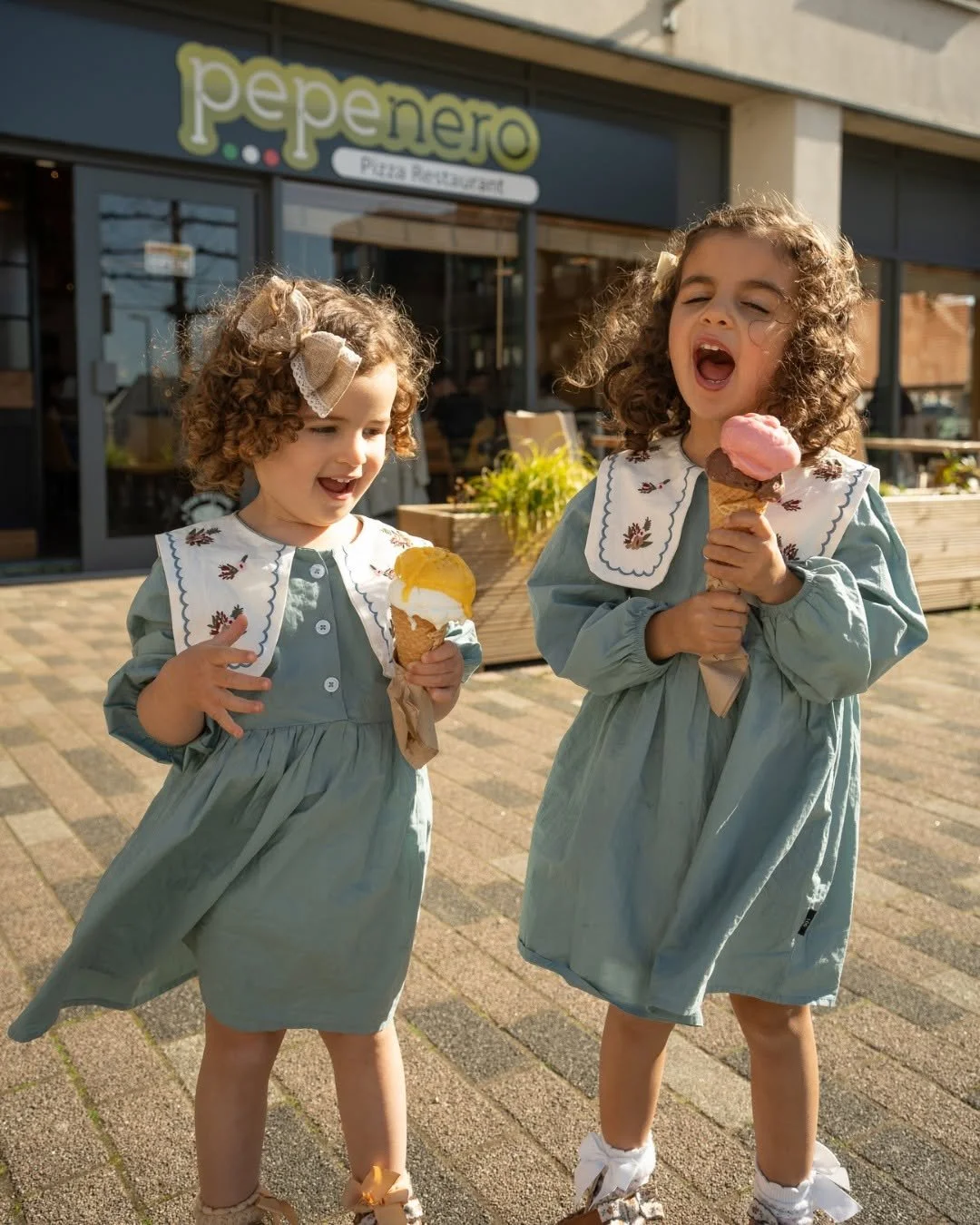Two young girls with curly hair wearing matching dresses and bows, each holding an ice cream cone, standing outdoors in front of a pizza restaurant called Pepe Nero.