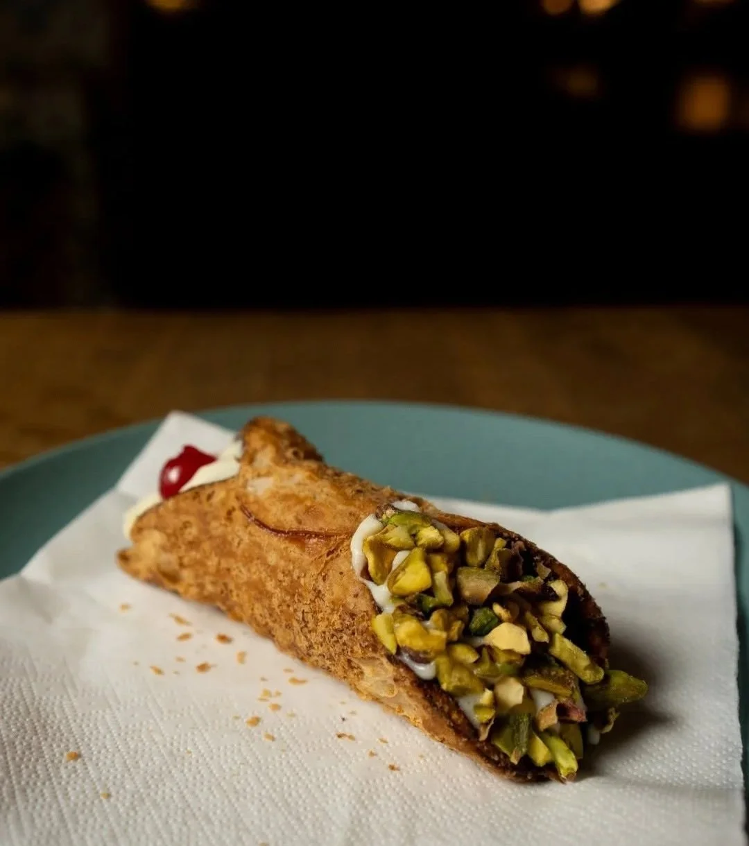 Pistachio cannoli with crushed pistachios and a cherry on top, served on a paper towel on a plate. PepeNero Pizza Restaurant