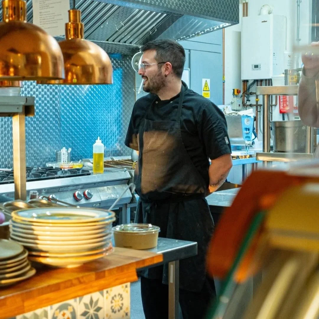 A male chef with glasses and dark hair in a black uniform and apron standing in a stainless steel kitchen. PepeNero Pizza Restaurant