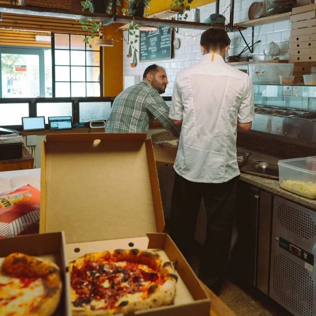Interior of a pizza restaurant with a customer and a staff member, nearby a pizza box with two slices of pizza on a table.