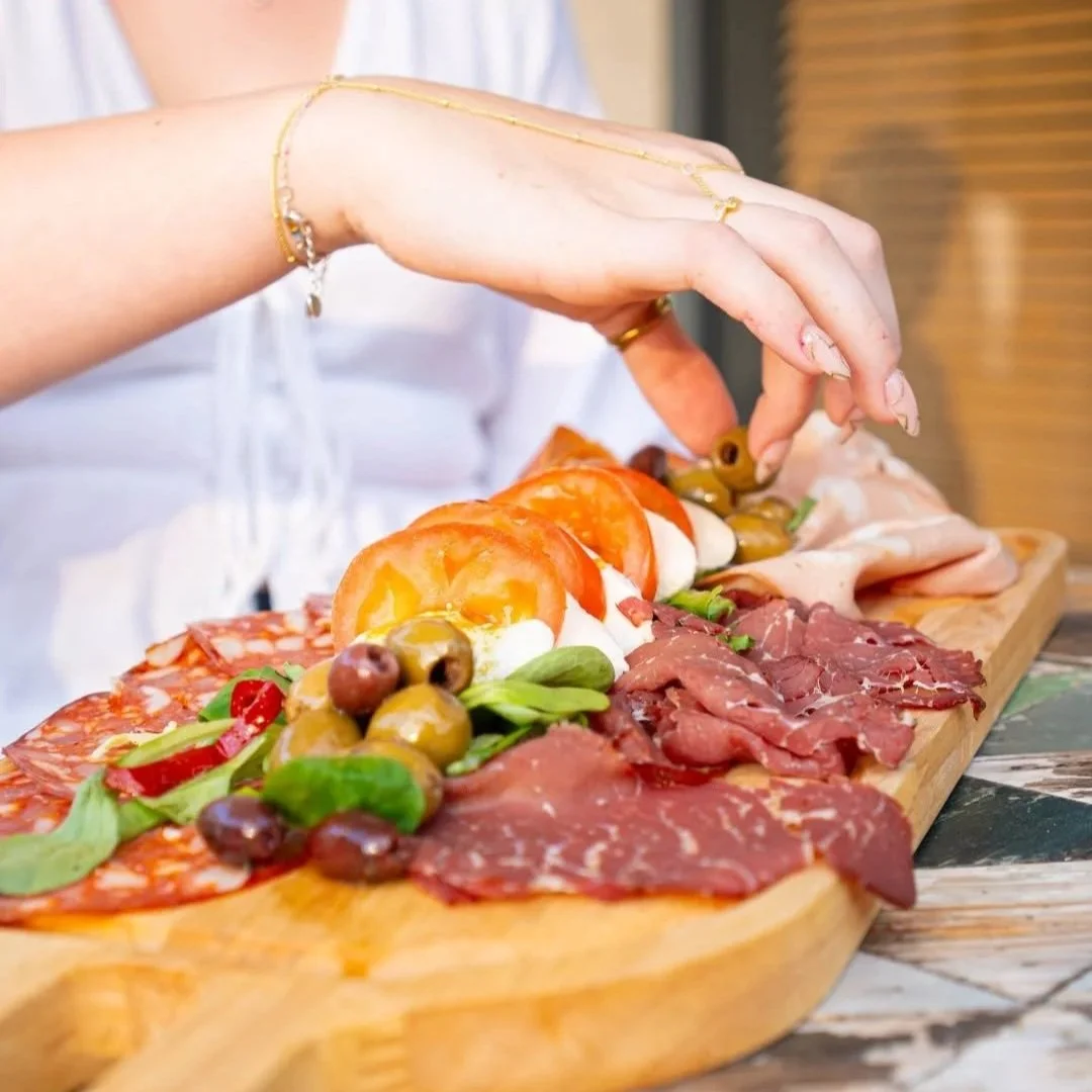 A hand reaching for sliced meats and cheese on a wooden charcuterie board with olives and vegetables.