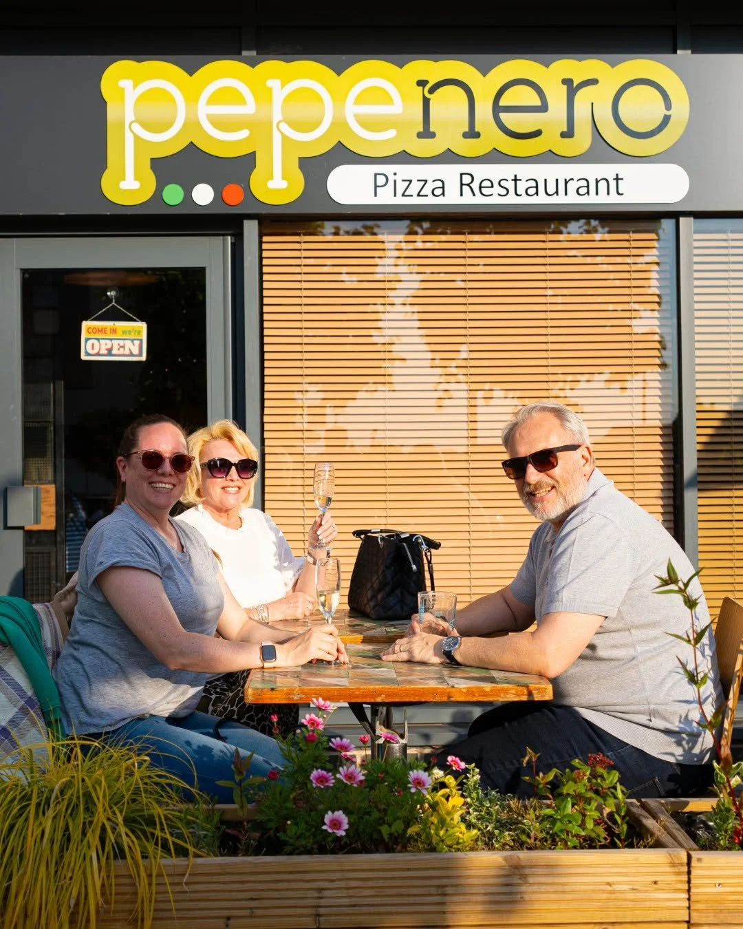 Four people sitting outside at a table in front of a pizza restaurant named Pepe Nero, smiling and holding glasses of champagne, with flowers in the foreground, and wearing sunglasses. PepeNero Pizza Restaurant