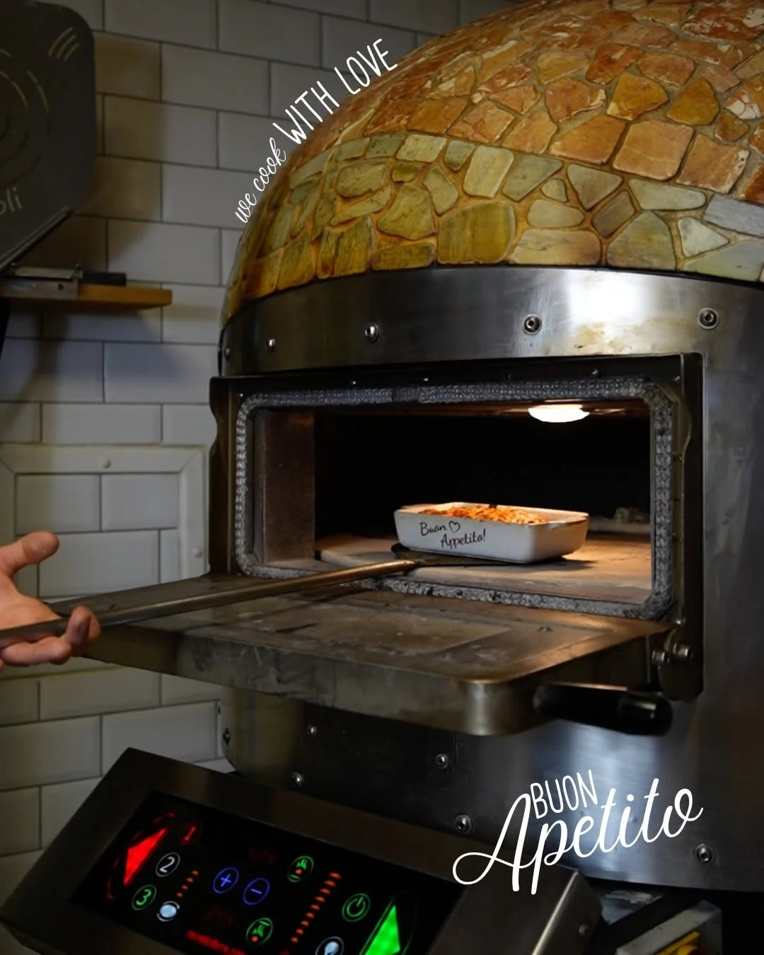 A pizza is being baked in a wood-fired oven with a stone exterior. The message on the oven window that says, "Buon Appetito!" and a tray inside the oven. Text on the image reads, "we cook with love" and "Buon Appetito". PepeNero Pizza Restaurant