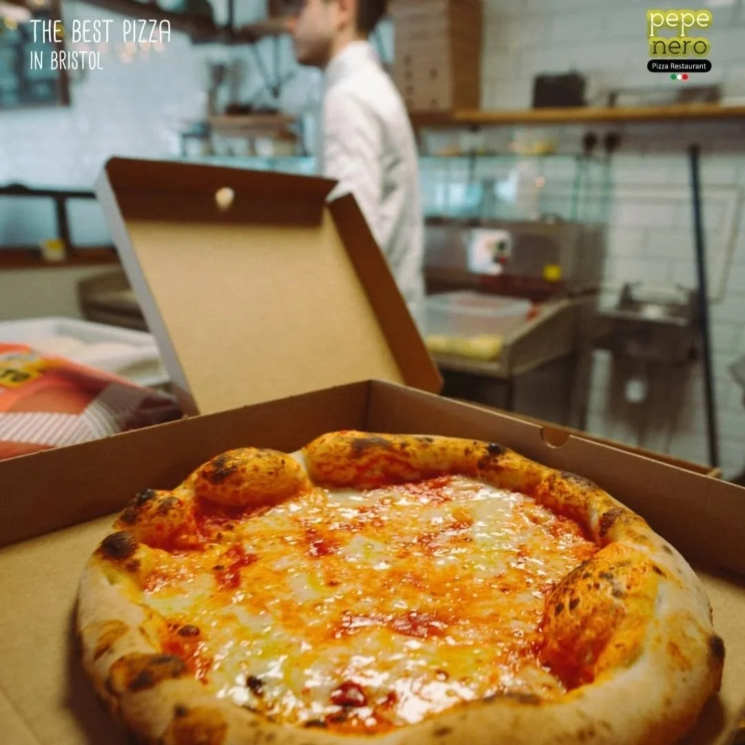 A freshly baked cheese pizza in an open pizza box with a restaurant setting in the background, including a blurry person and shelves.