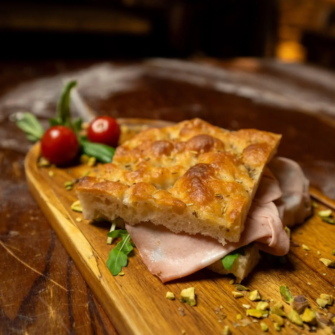A slice of quiche with a golden crust on a wooden serving board, garnished with cherry tomatoes and basil, with pieces of chopped pistachios and some arugula underneath. PepeNero Pizza Restaurant