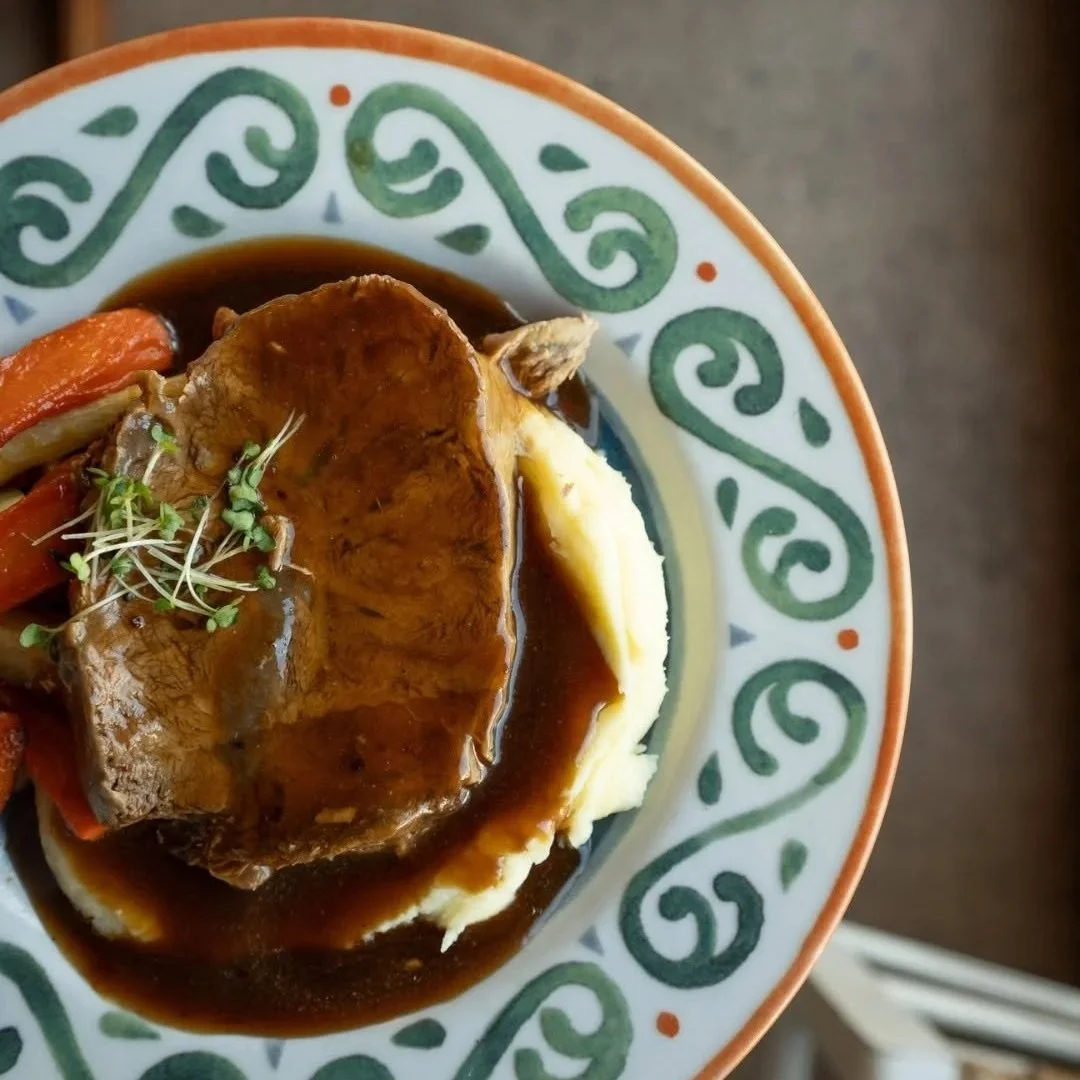 A bowl of mashed potatoes topped with beef stew, carrots, and microgreens, served with brown gravy in a decorative patterned bowl.