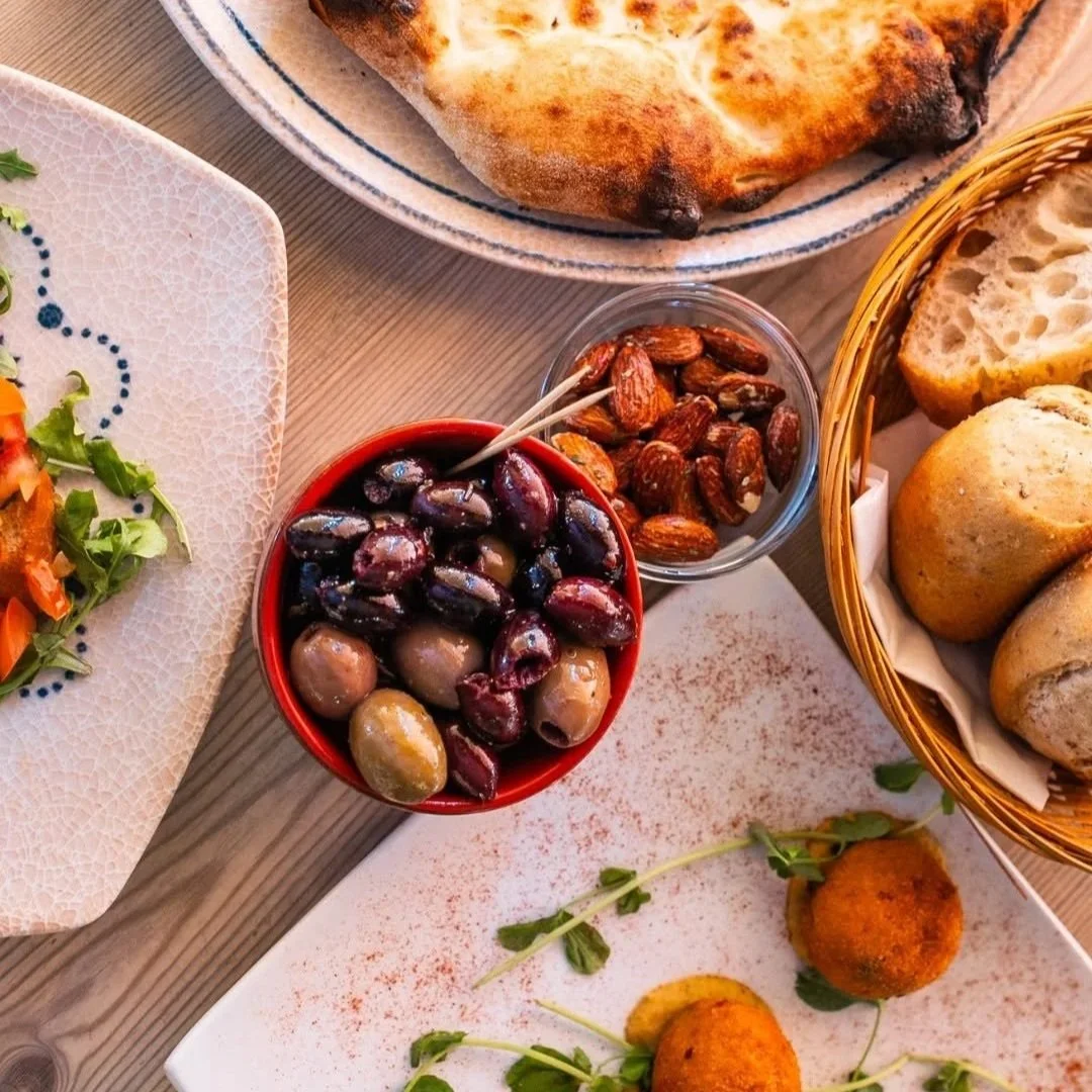 Assorted Mediterranean dishes including a bowl of mixed olives, a basket of bread rolls, toasted pita bread, baked eggplant, and a salad with vegetables. PepeNero Pizza Restaurant