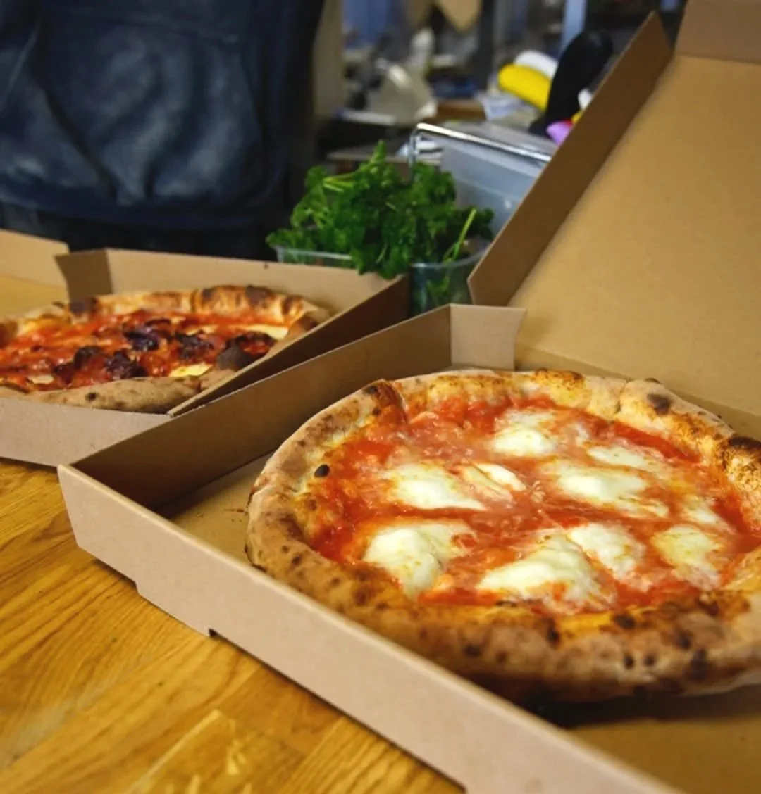 Two open pizza boxes, one with a Margherita pizza and the other with a pizza topped with pepperoni and sausage, on a wooden table. PepeNero Pizza Restaurant