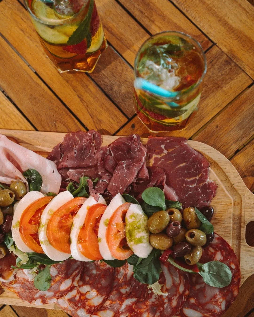 Charcuterie board with sliced cured meats, fresh mozzarella, tomatoes, green and black olives, and greens on a wooden serving board, with two glasses of iced tea on a wooden table.