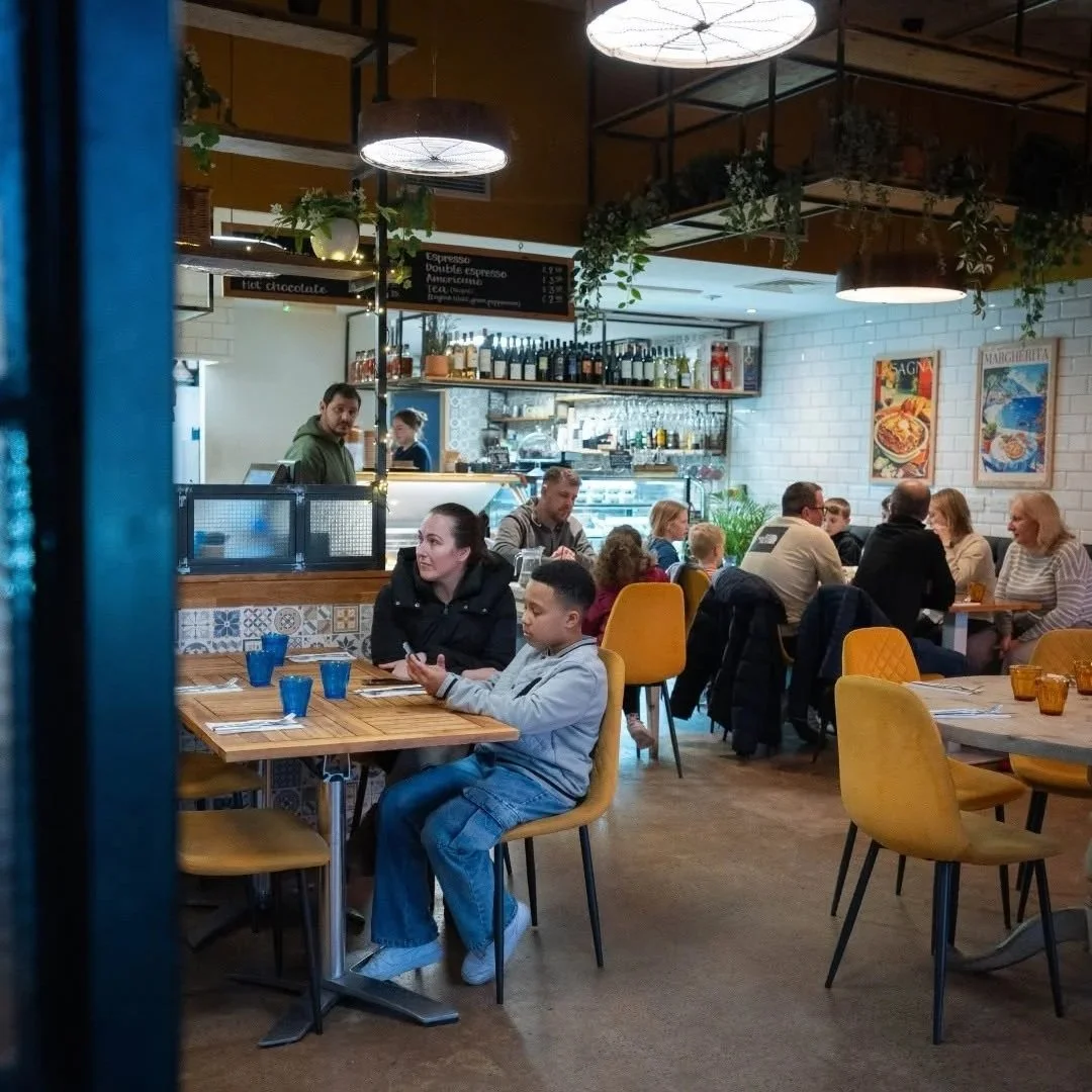 Interior of a lively restaurant with patrons seated at tables, a visible bar area, and hanging ceiling lights, with blue and amber glassware on the tables.