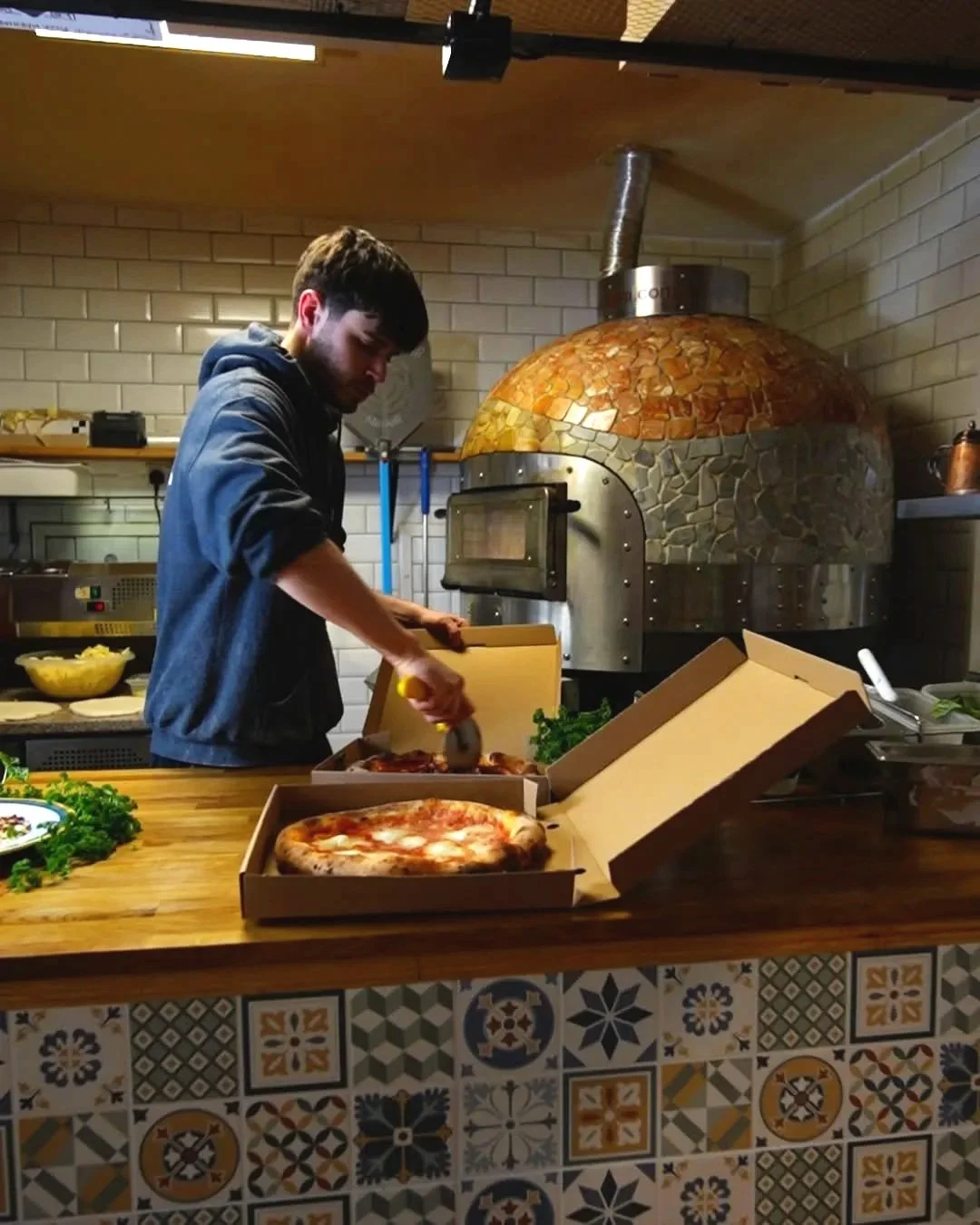 A man cutting a pizza in a restaurant kitchen with a wood-fired oven in the background.