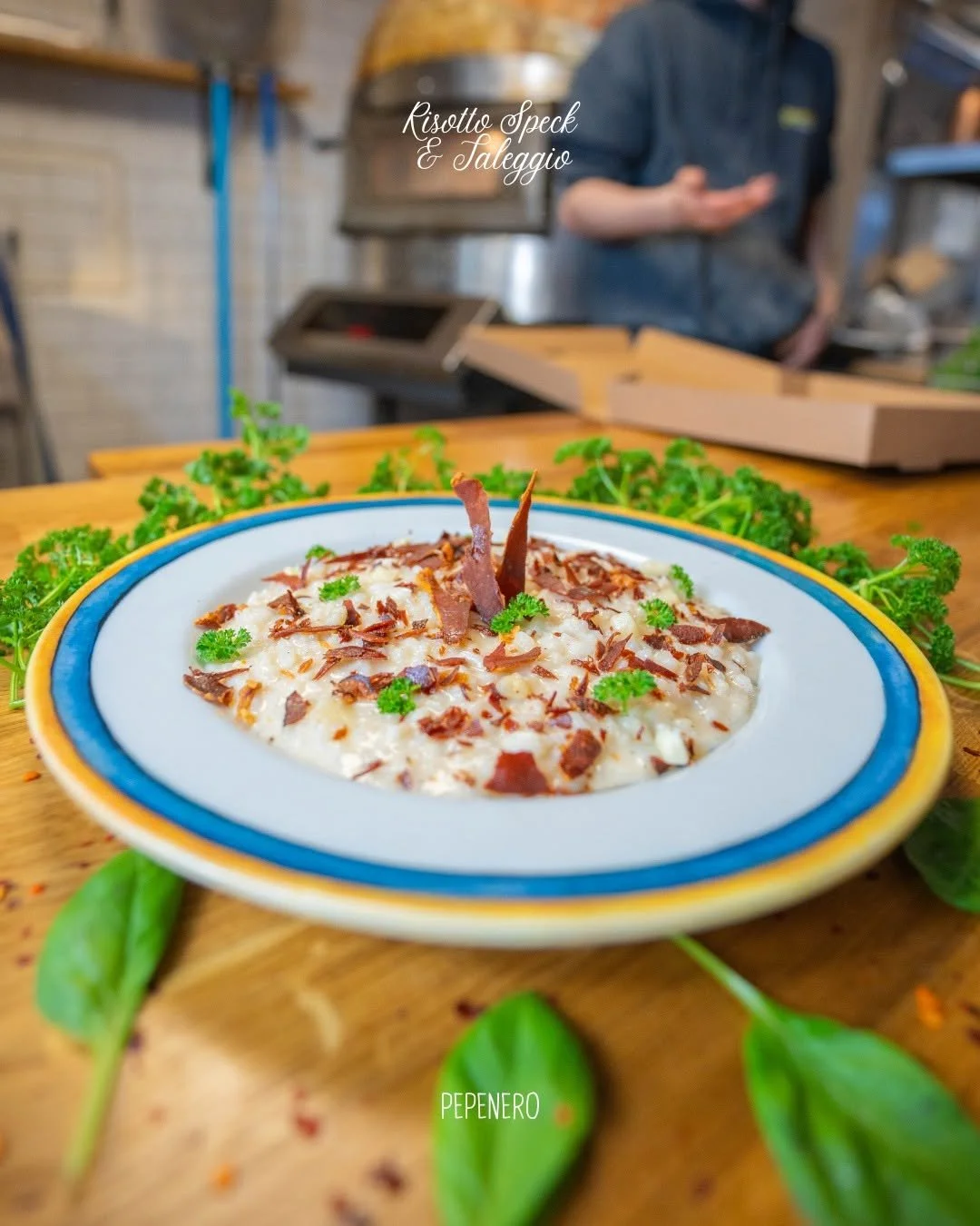 A plate of risotto speck with parsley, garnished with parsley leaves, on a wooden table surrounded by green herbs, with a chef in the background in a kitchen setting. PepeNero Pizza Restaurant