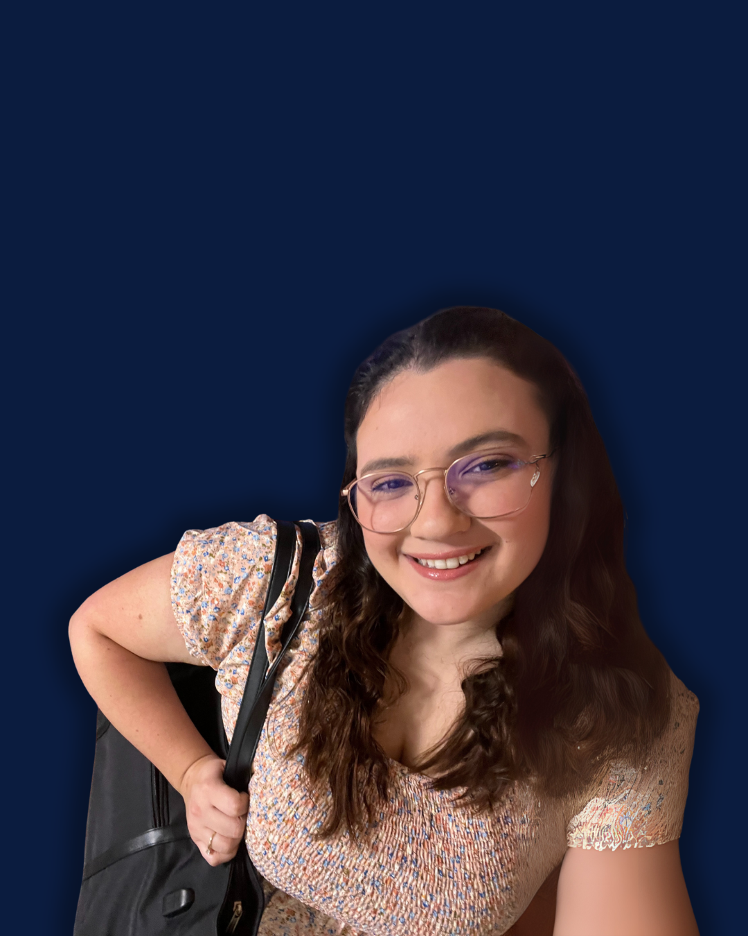 A young woman with glasses and long curly hair smiling at the camera, wearing a floral dress and carrying a black backpack over her shoulder against a dark blue background.