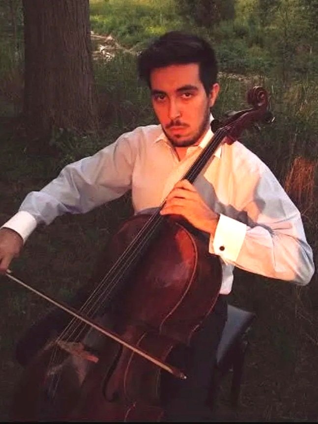 A young man with dark hair and a beard, wearing a white dress shirt with black buttons, is playing a cello outdoors near a tree.