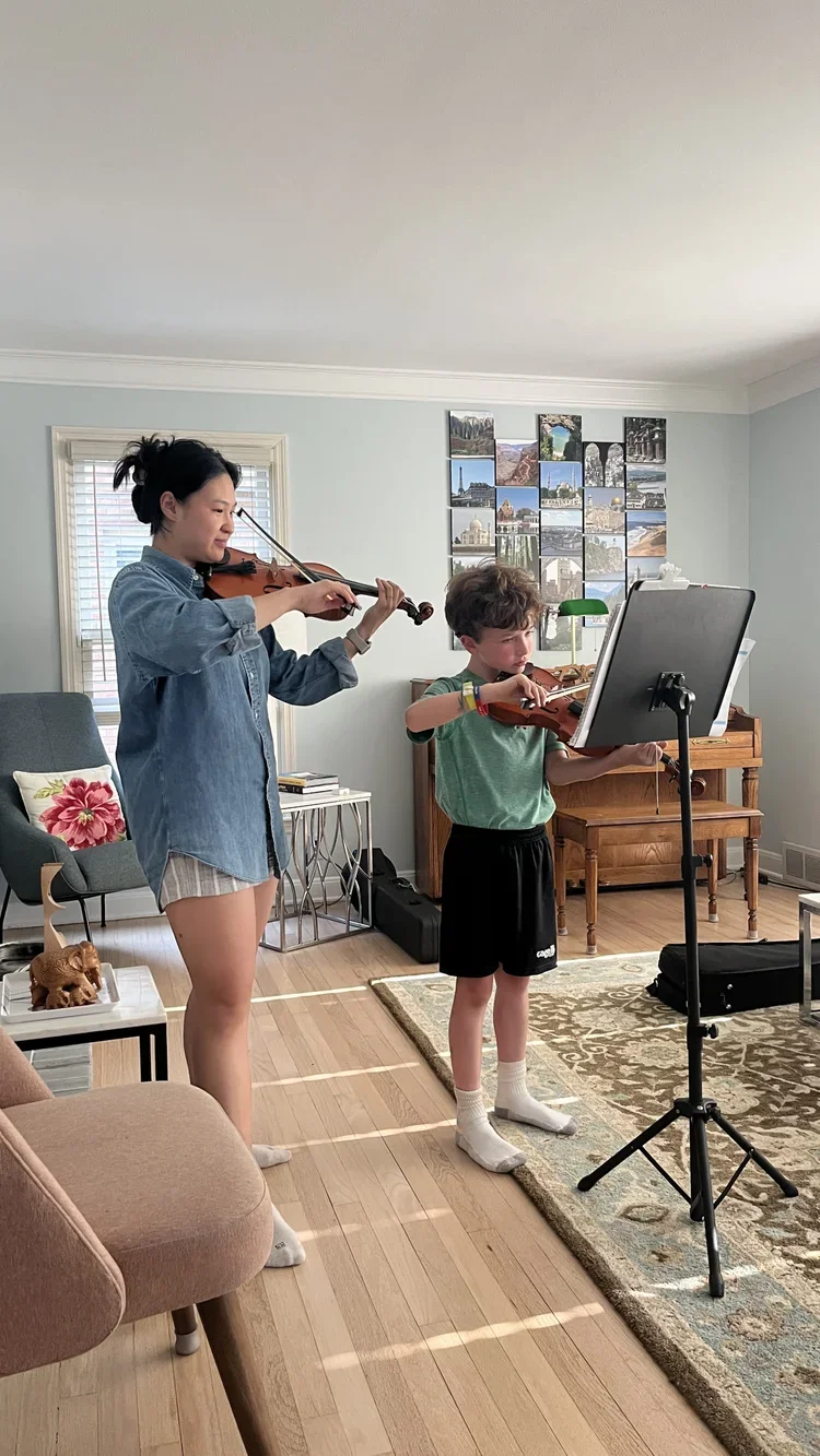 A woman and a young boy are playing violins in a living room. The woman is standing and the boy is standing at a music stand. The room has a light-colored wall, a window with blinds, a framed photo collage, and various furniture including a chair with a floral pillow, a small table, and a wooden piano. There is a dog figurine on a small table and a rug on the wooden floor.