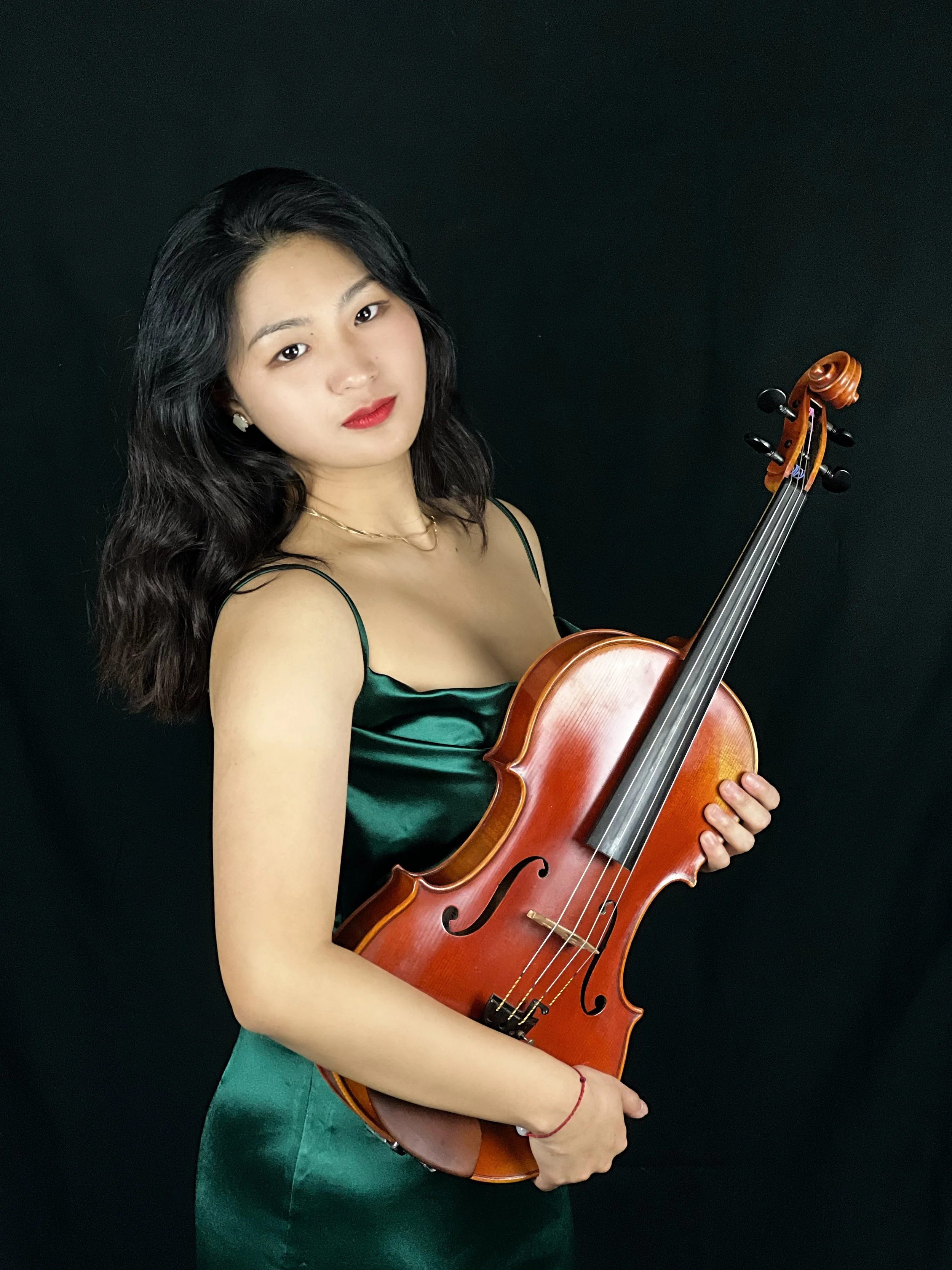 A woman with long dark hair in a green satin dress holding a violin against a black background.