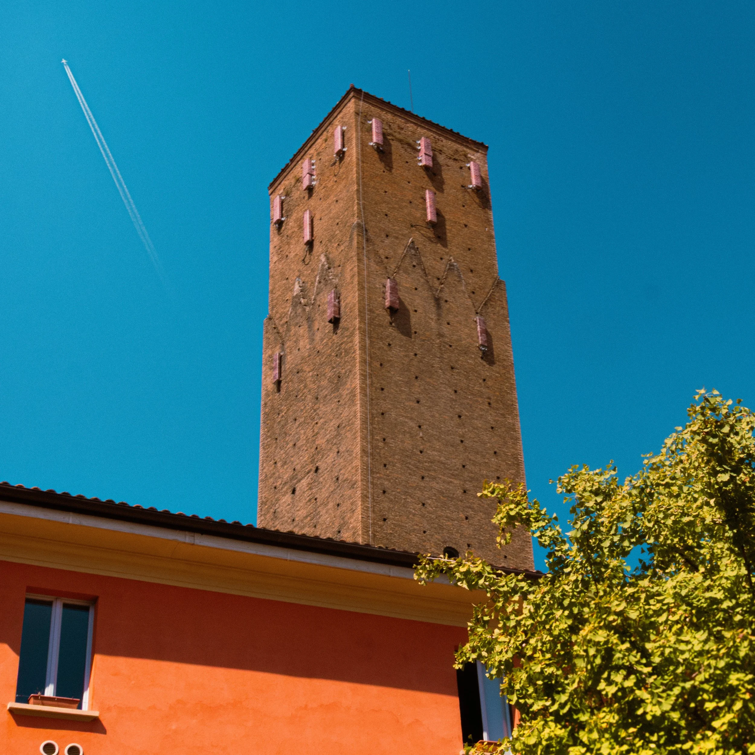 A tall brick tower with pink fixtures on the facade, partly obscured by a tree and a building with a red roof, under a clear blue sky with contrails.
