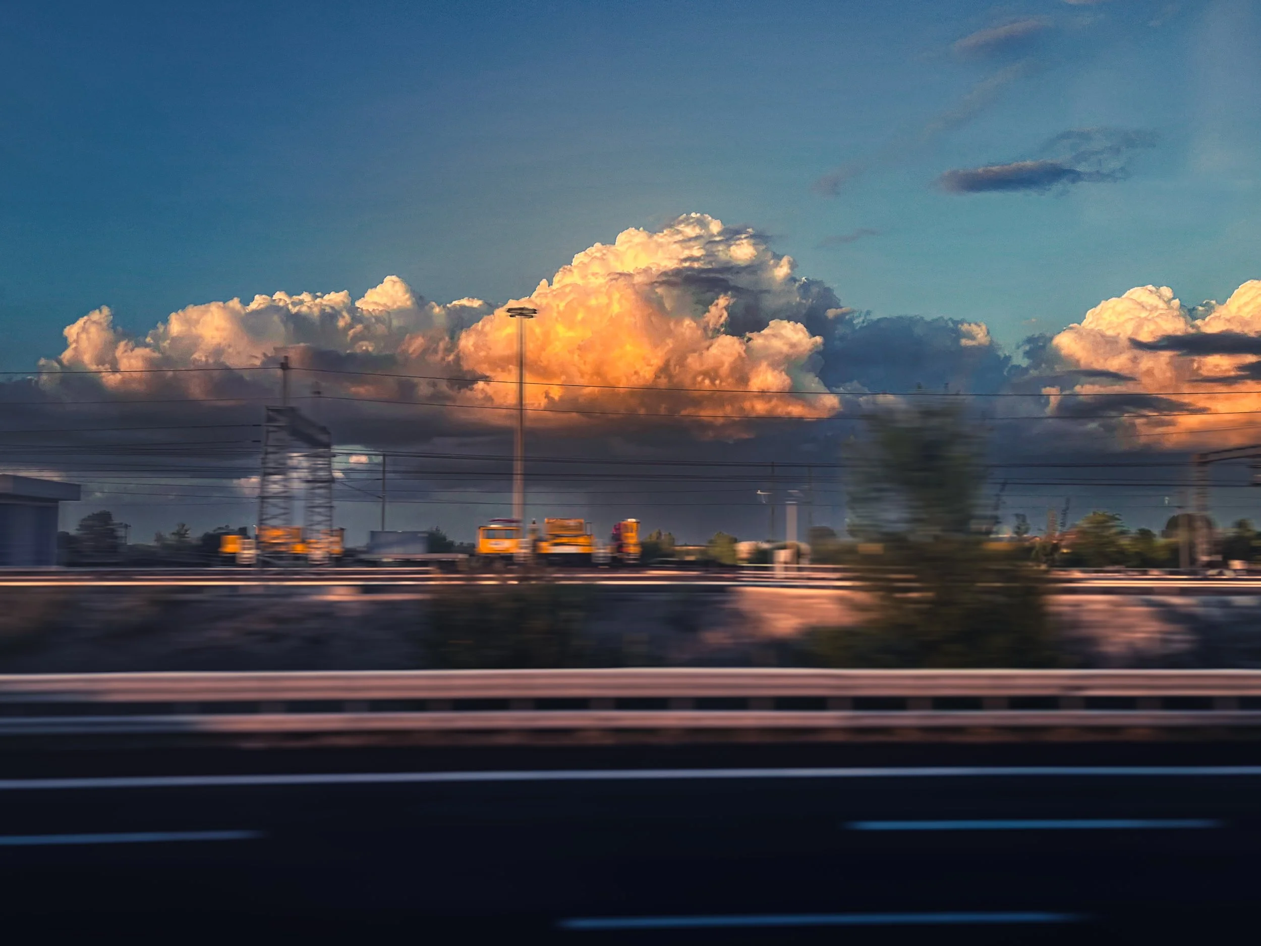 A highway with a guardrail in the foreground and a cityscape in the background under a sky with large orange and yellow cloud formations at sunset.