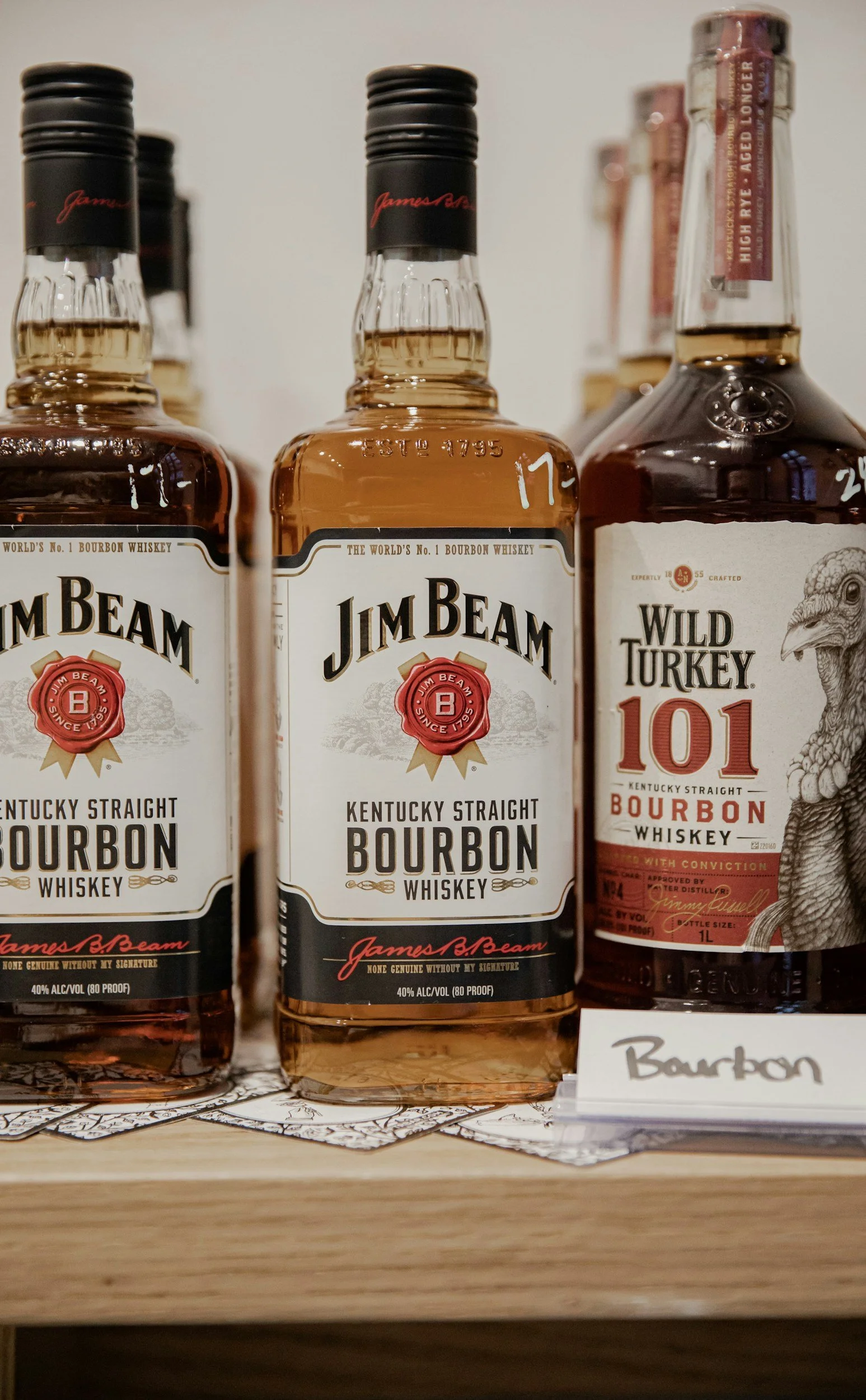 Three bottles of bourbon whiskey on a wooden surface, with two Jim Beam Kentucky Straight Bourbon bottles in the front and a Wild Turkey 101 bottle to the right. There are small sheets of paper with handwritten labels in front of the bottles.
