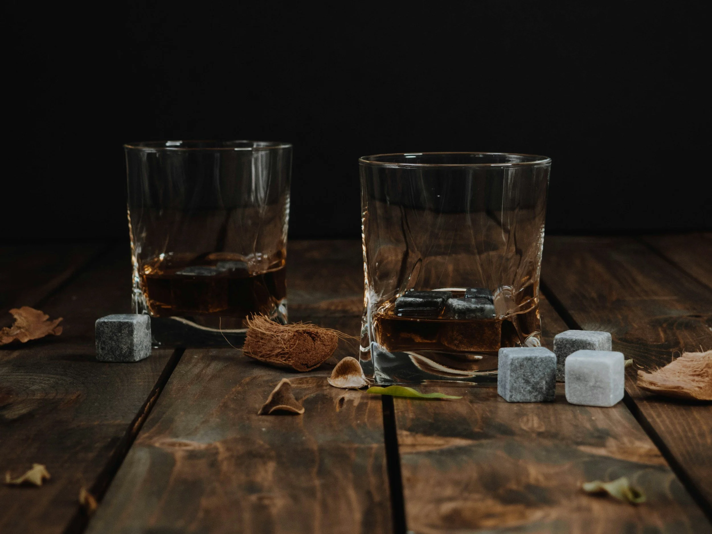 Two glasses of whiskey on a wooden table surrounded by scattered leaves, ice cubes, and small stones.