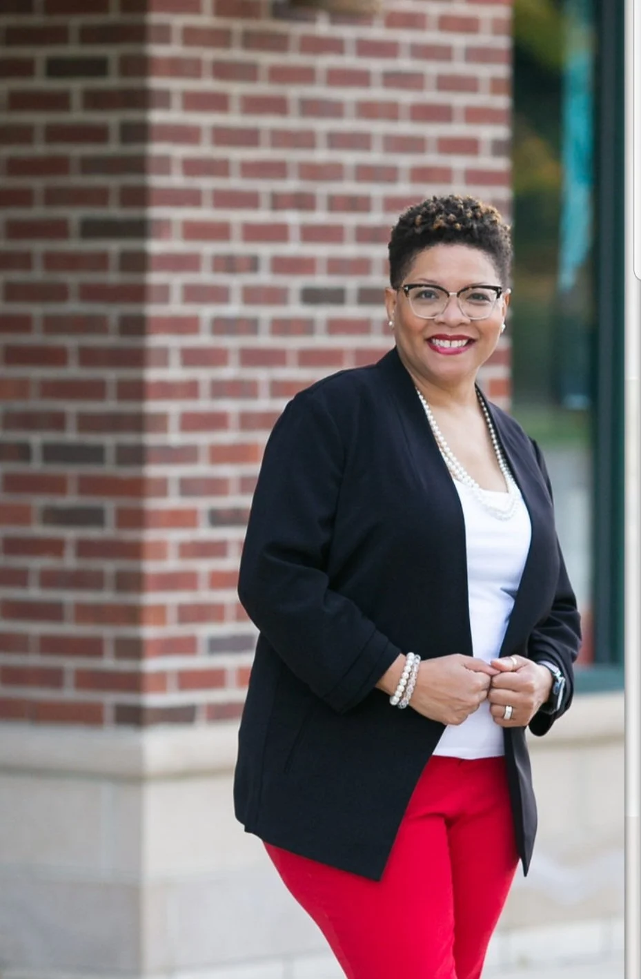 A woman with short curly hair, glasses, and a wide smile, standing outside against a brick wall, wearing a black blazer, white top, red pants, and accessorized with pearl jewelry.