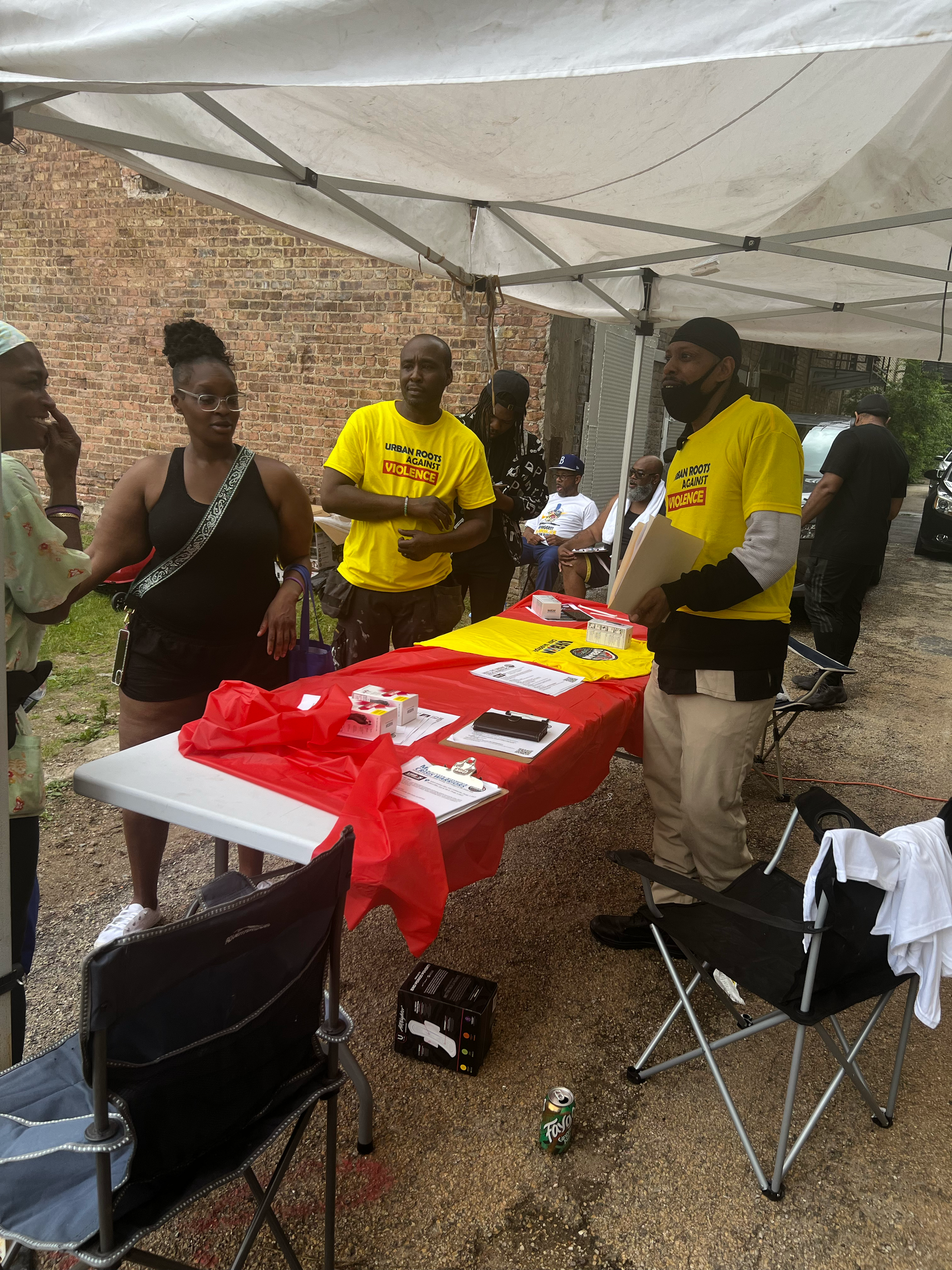 People gathered under a tent at an outdoor event, some wearing yellow shirts with a message about ending violence against women, with tables displaying pamphlets and materials.