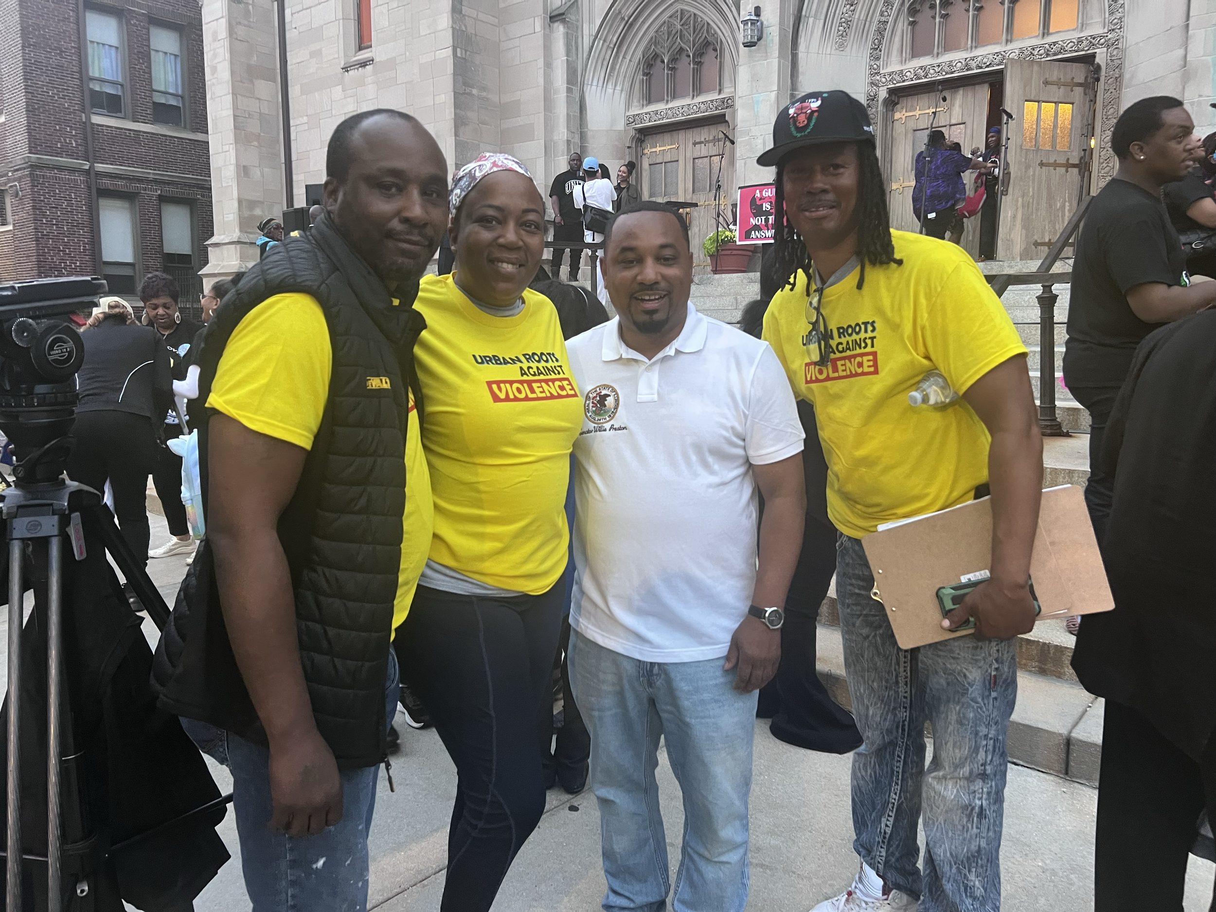 Four people standing together outside a church, two men and two women, at a protest or rally against violence. The women and one man are wearing yellow shirts with the text 'Urban Roots Against Violence.' The other man is wearing a white polo shirt.