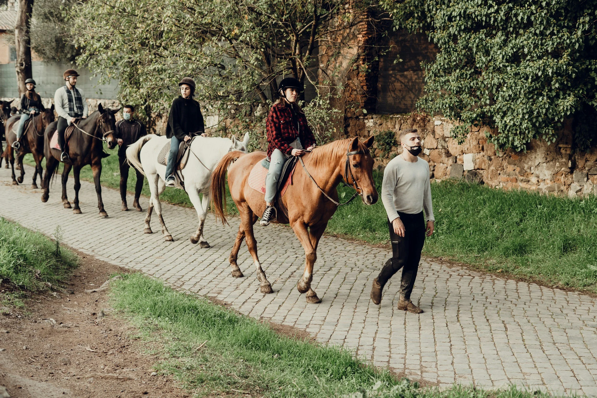 People riding horses on a cobblestone path outdoors with greenery and a stone wall in the background.