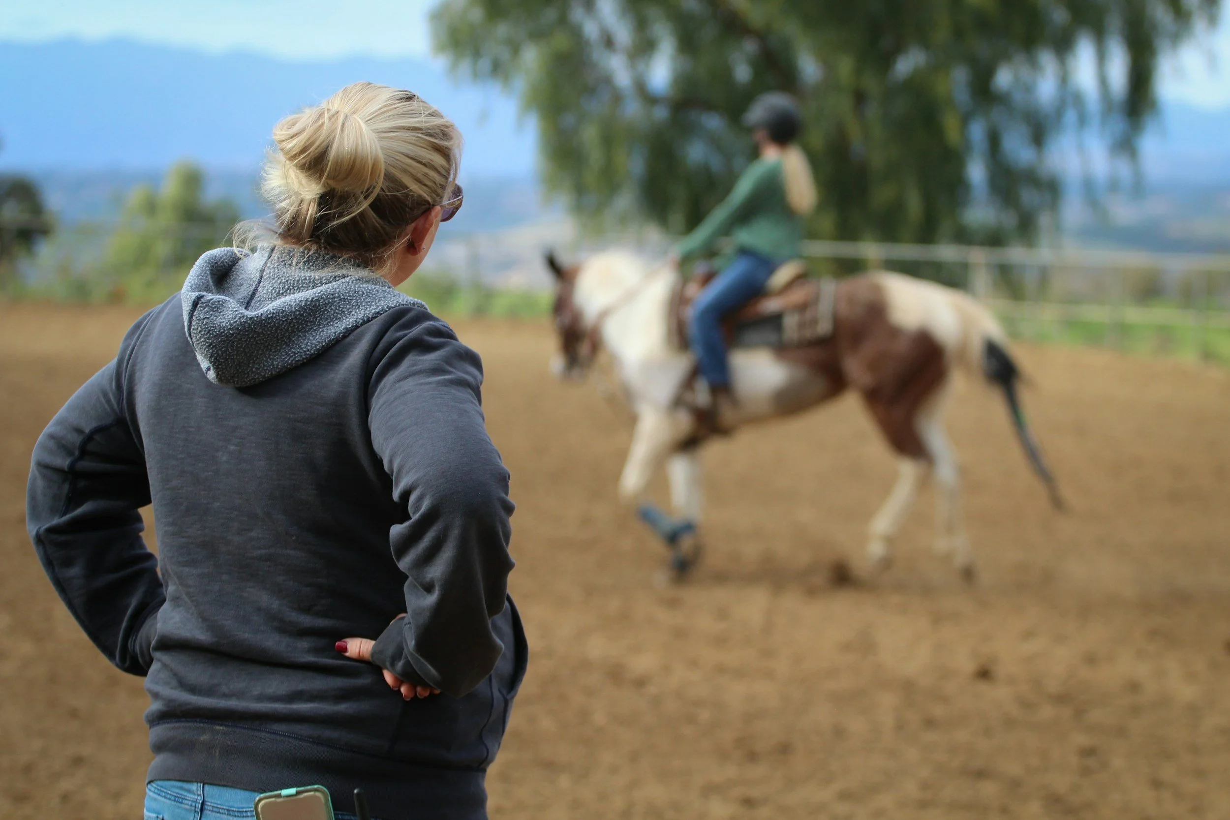 A woman with blonde hair in a bun, wearing glasses and a black hooded jacket, watches a girl riding a horse in an outdoor riding arena. The girl is wearing a helmet and a green sweater, and is riding a brown and white horse. The background shows trees and mountains.