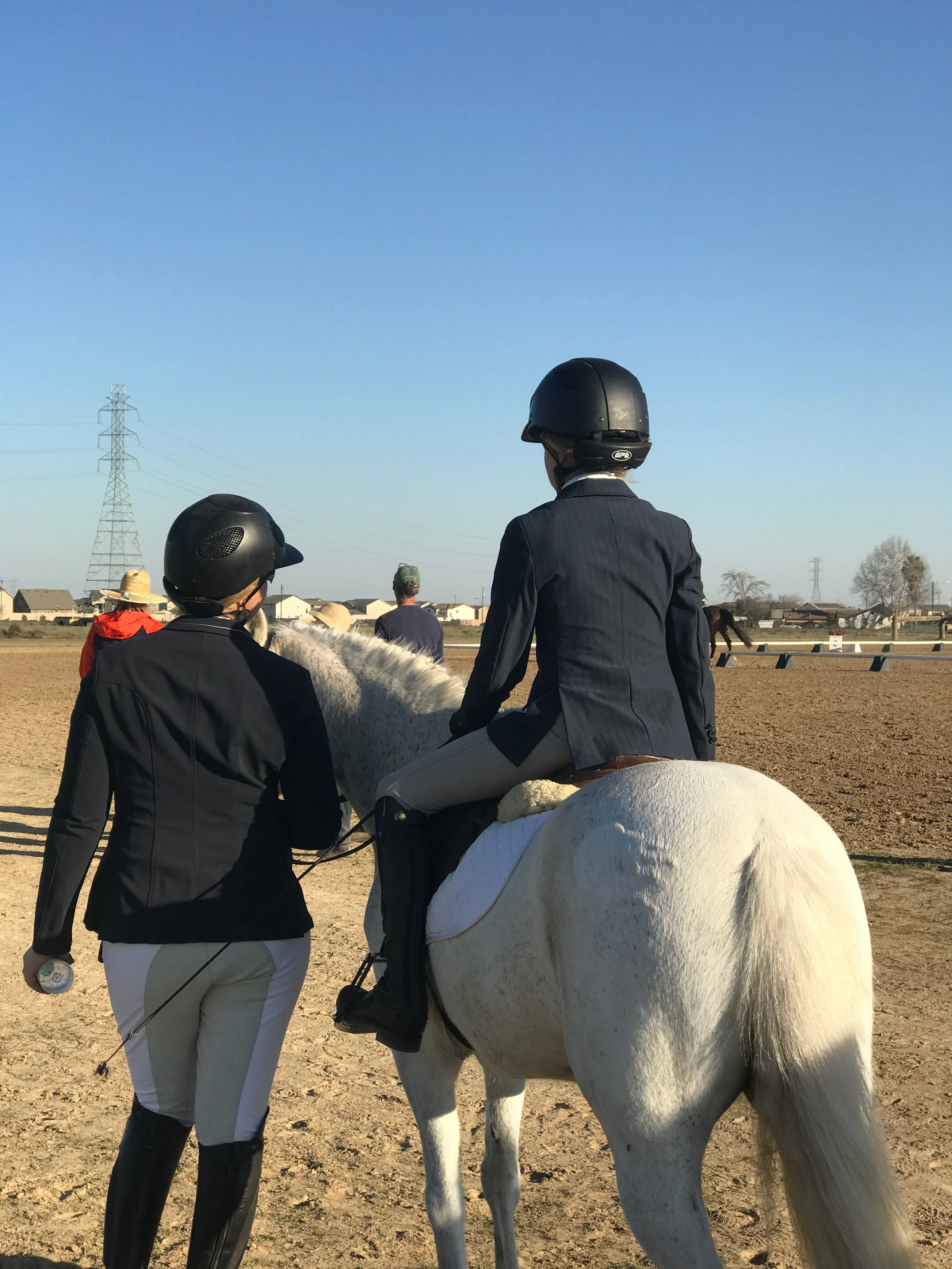 Child on a white pony with an instructor walking beside them at a horse show.