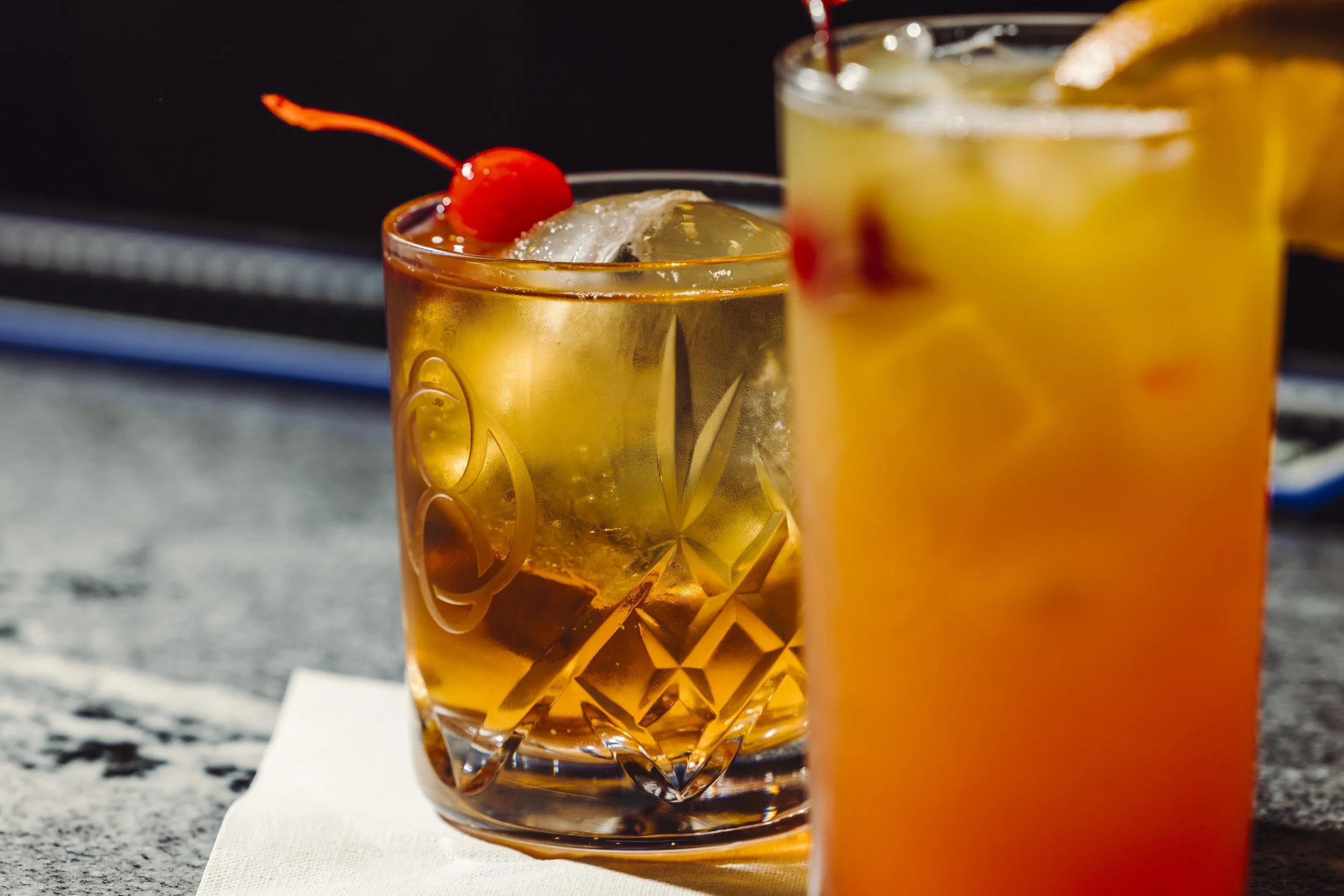 Two cocktails, one with a maraschino cherry on top, on a surface with a white napkin.