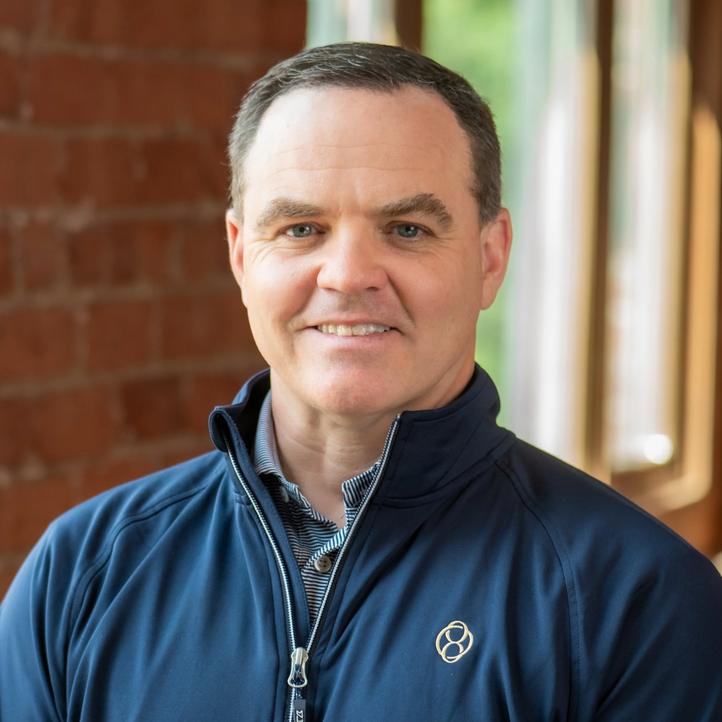 Close-up of a smiling man wearing a black zip-up jacket with a gold embroidered logo on the chest, standing indoors near a window with green foliage outside.