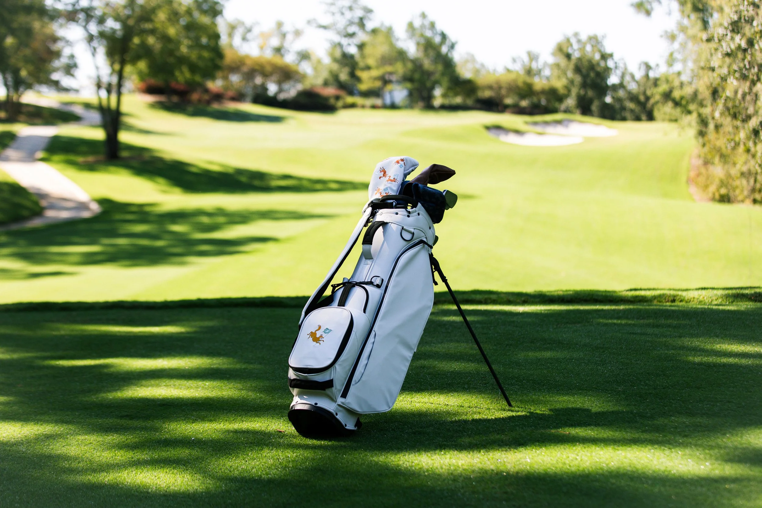 A golf bag with clubs stands on a lush green golf course with trees and sand traps in the background.