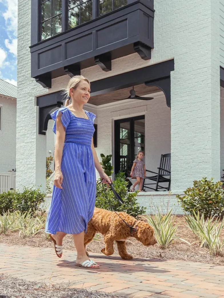 A woman in a blue striped dress walking a brown dog on a leash in front of a modern house with a white brick exterior and a covered porch. A young girl in a floral dress is standing on the porch.