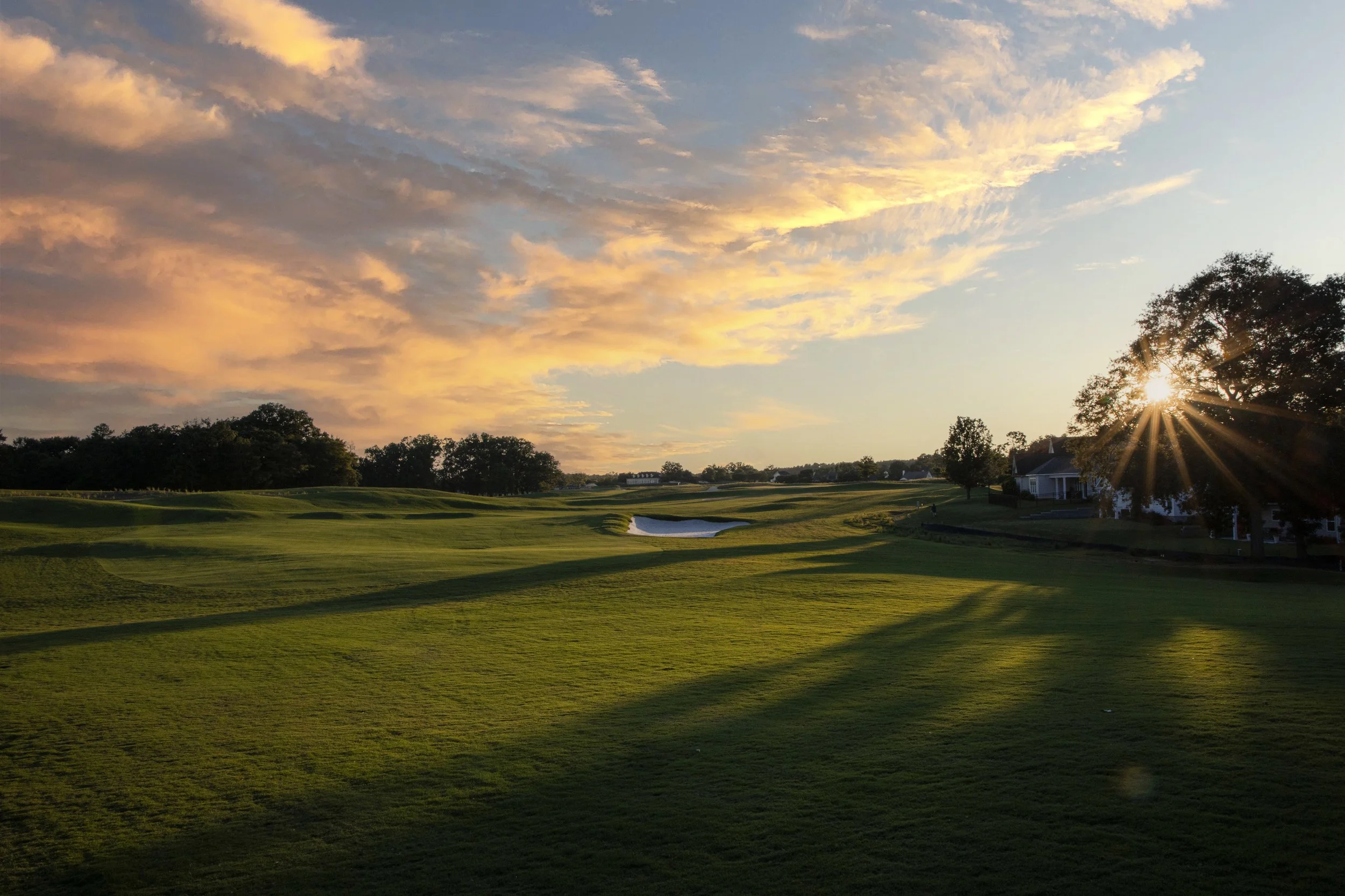 Sunset view over a golf course with green grass, sand trap, trees, and a house with the sun shining through a tree.