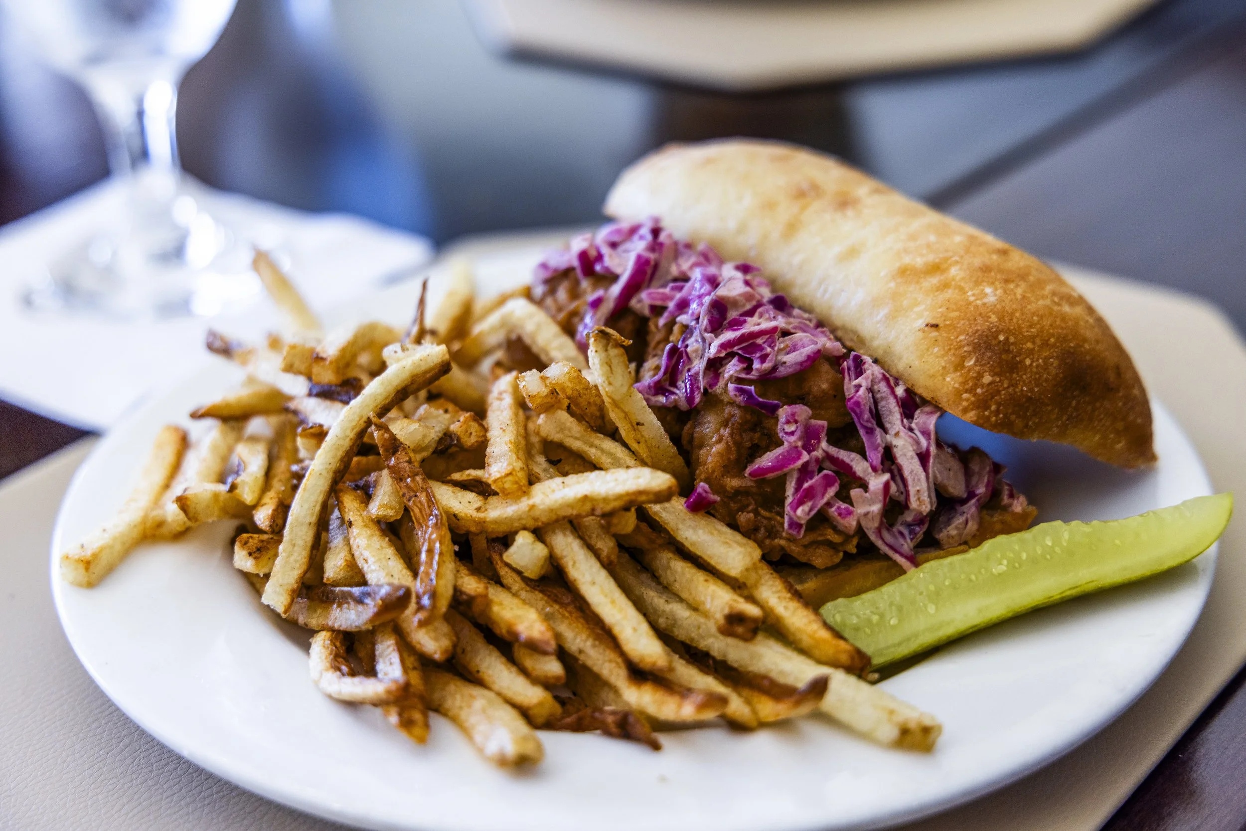 French bread sandwich filled with pulled pork and purple coleslaw, served with French fries and pickle slices on a white plate.